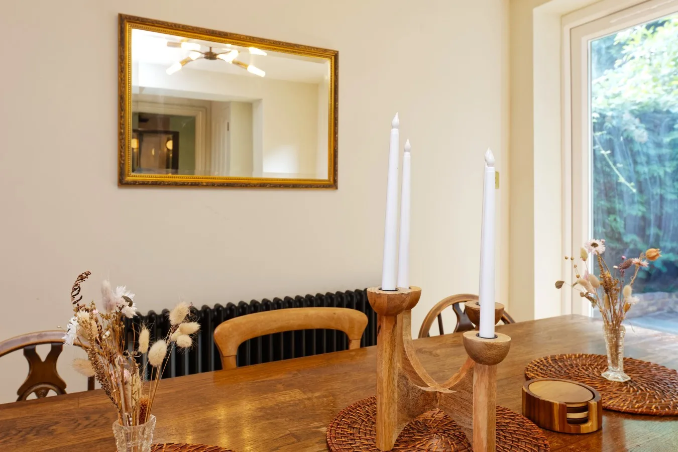 A wooden dining table with candles and dried flowers sits beside a large window.