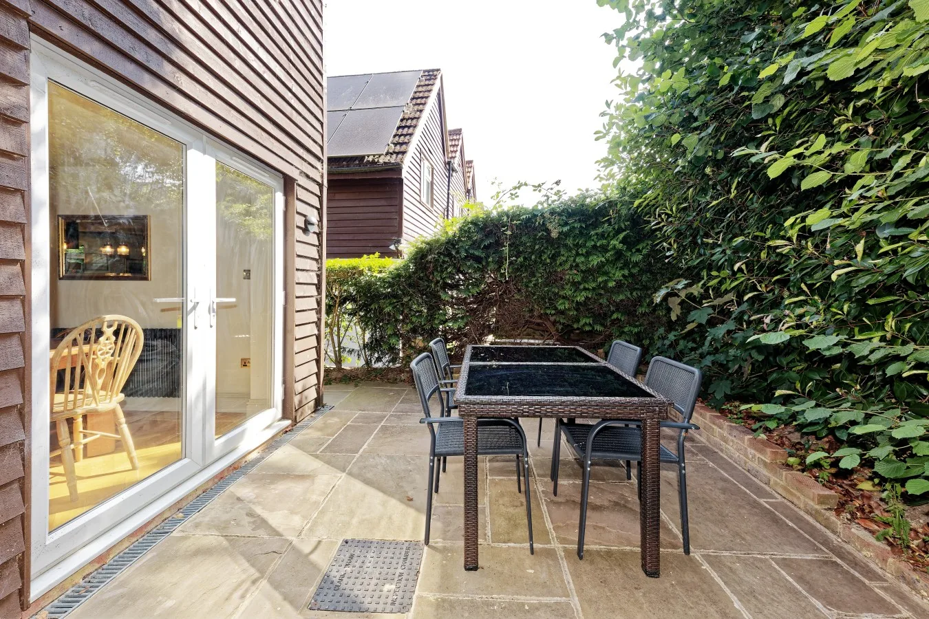 A paved patio with an outdoor dining table and chairs beside a hedge.