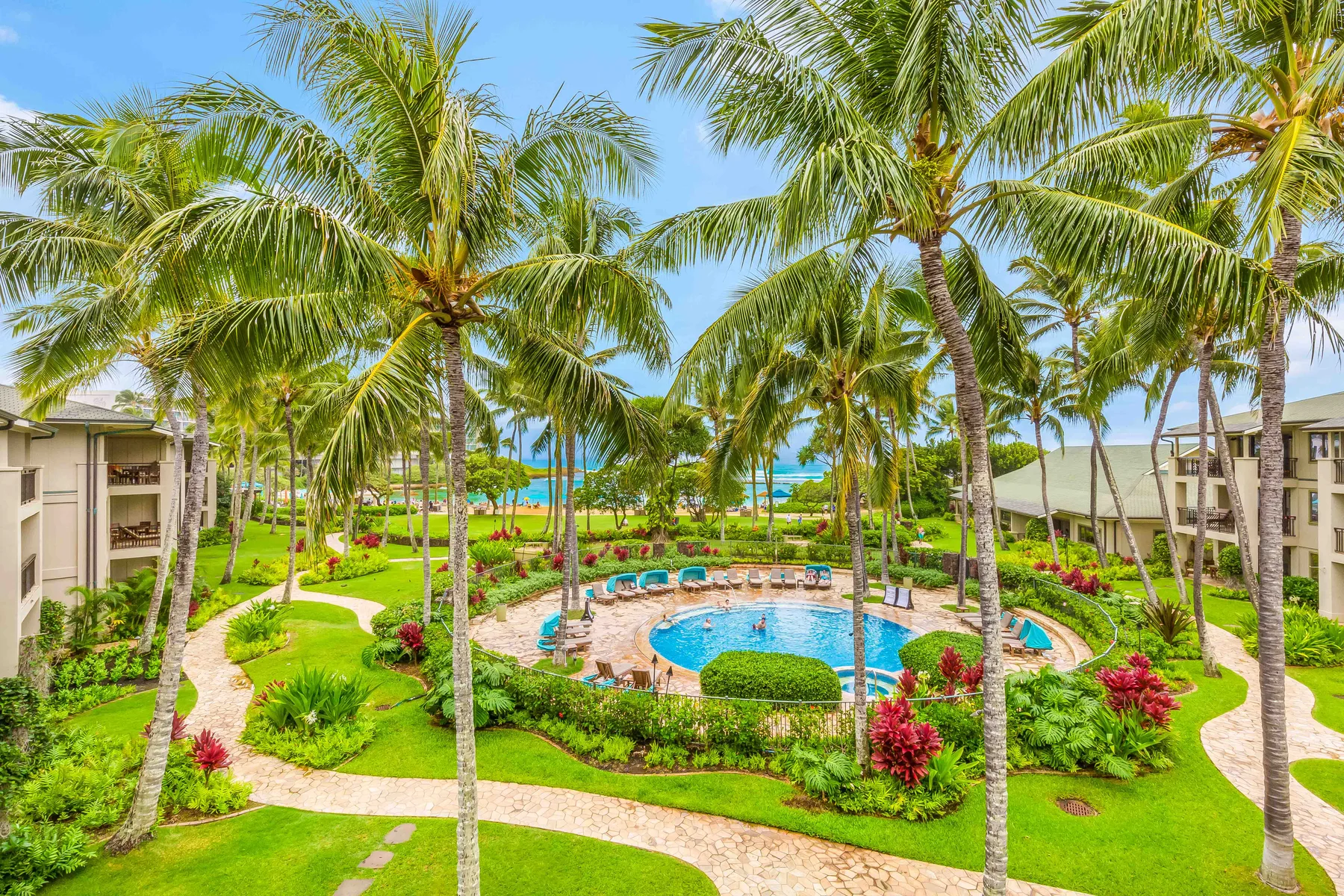 Zona de piscina ajardinada con palmeras, jardines y senderos curvos.