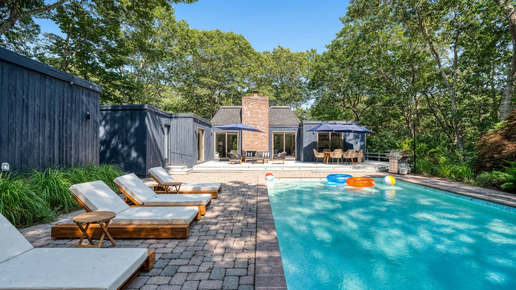 Una piscina al aire libre junto a un patio pavimentado, tumbonas y árboles.