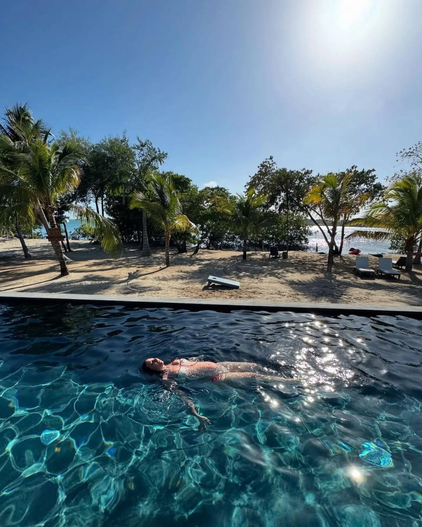 Una persona flota en una piscina con vistas a una playa bordeada de palmeras.