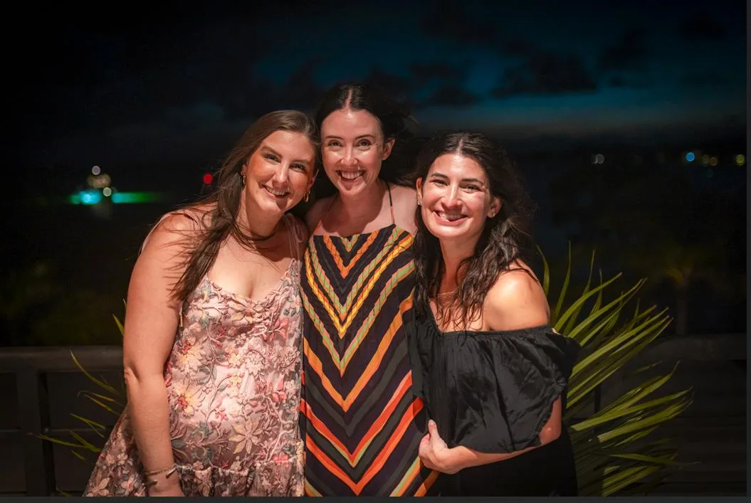 Tres mujeres posan juntas al aire libre por la noche junto a plantas frondosas.