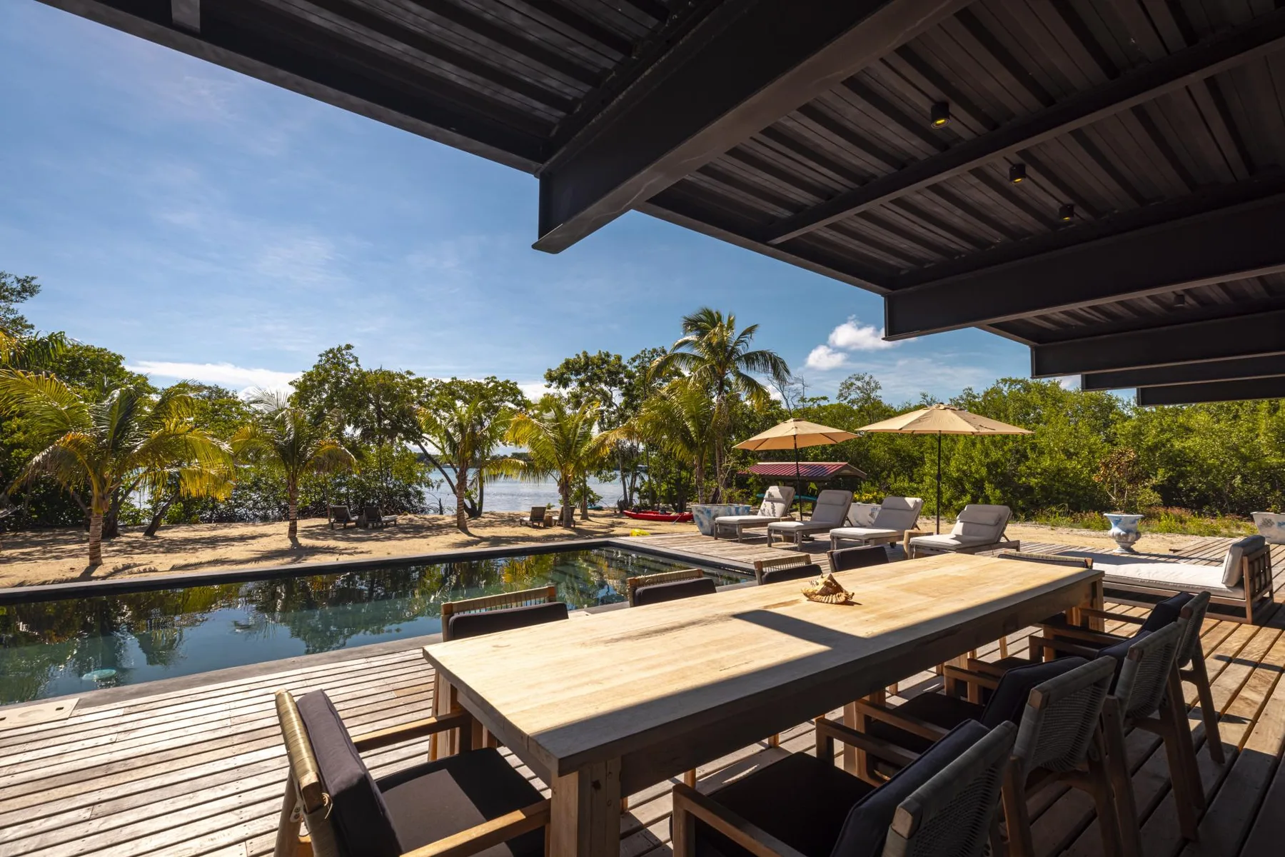 Un comedor al aire libre da a una piscina, con sombrillas y vegetación tropical al fondo.