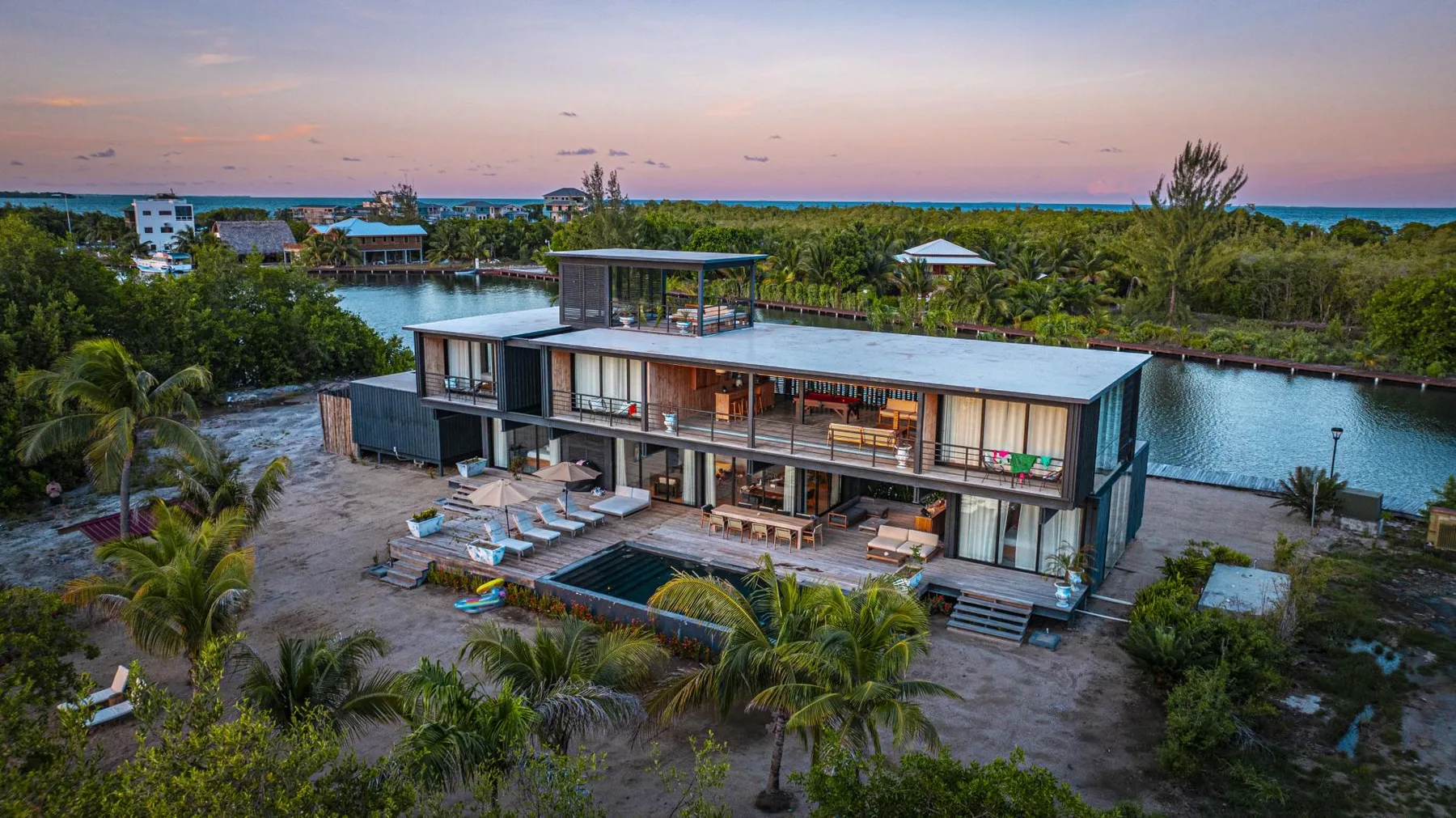 A modern house beside a waterway, surrounded by palm trees at dusk.