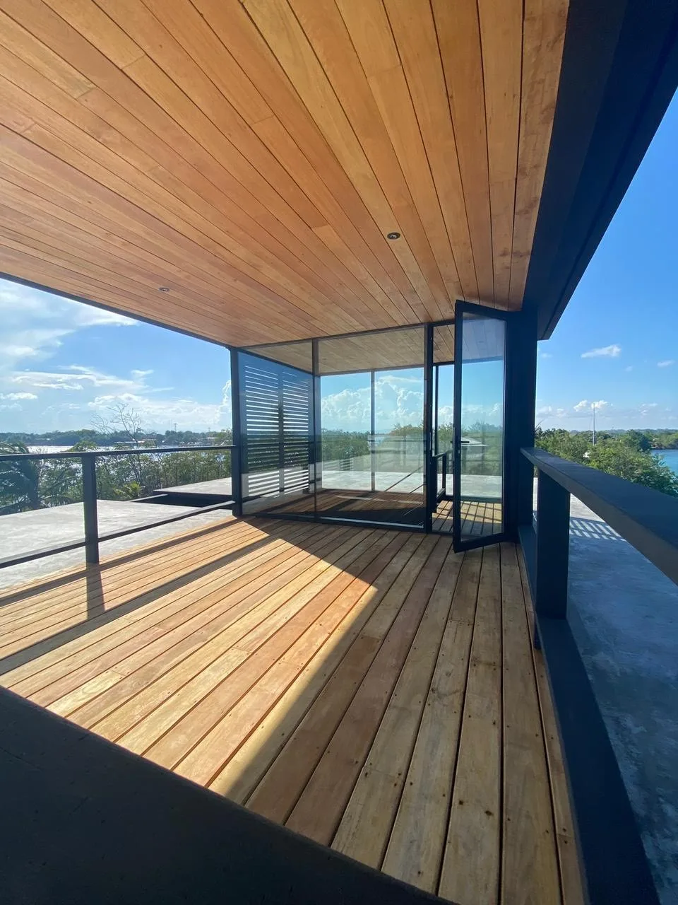 Una terraza de madera iluminada por el sol, con paredes de vidrio, da a un paisaje verde.