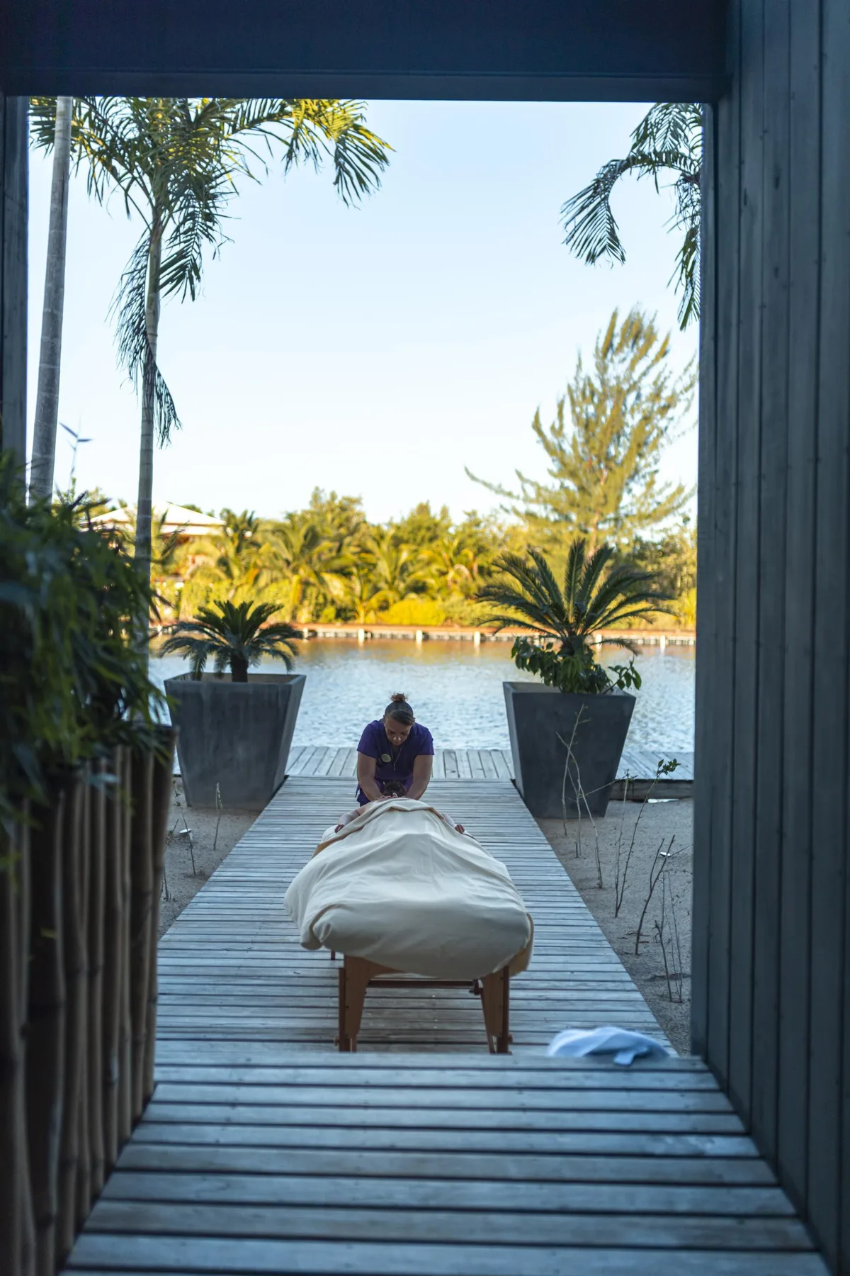 Una camilla de masaje está sobre una pasarela de madera con vistas a una piscina rodeada de palmeras.