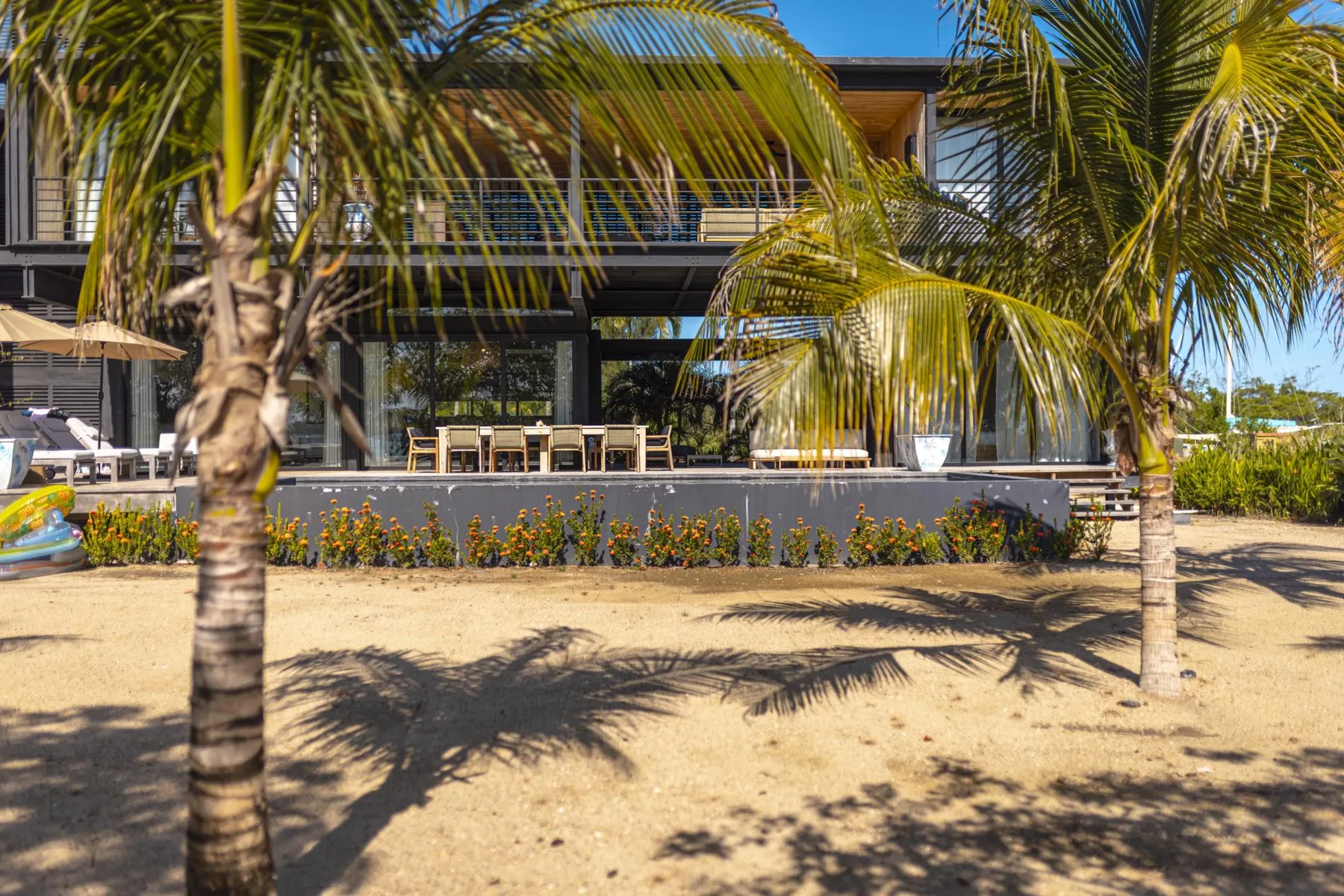 Un edificio moderno con una zona de asientos al aire libre a la sombra y palmeras.