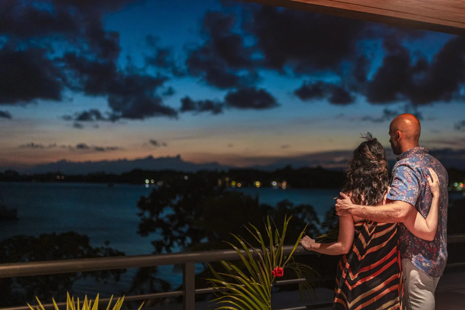 Una pareja está en un balcón con vistas al agua bajo un cielo nublado al atardecer.