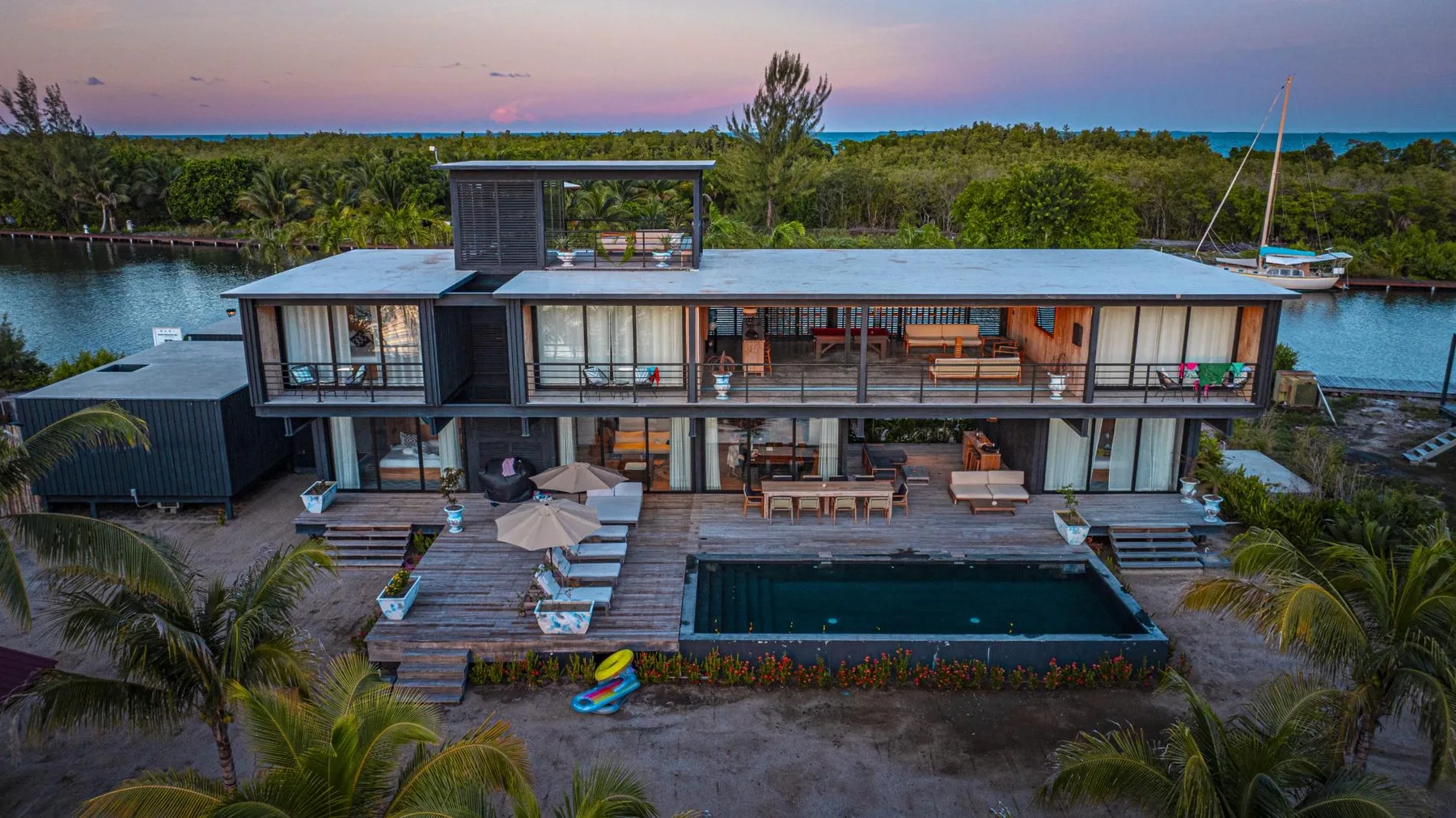 Una casa moderna junto al agua, con piscina y asientos al aire libre al anochecer.