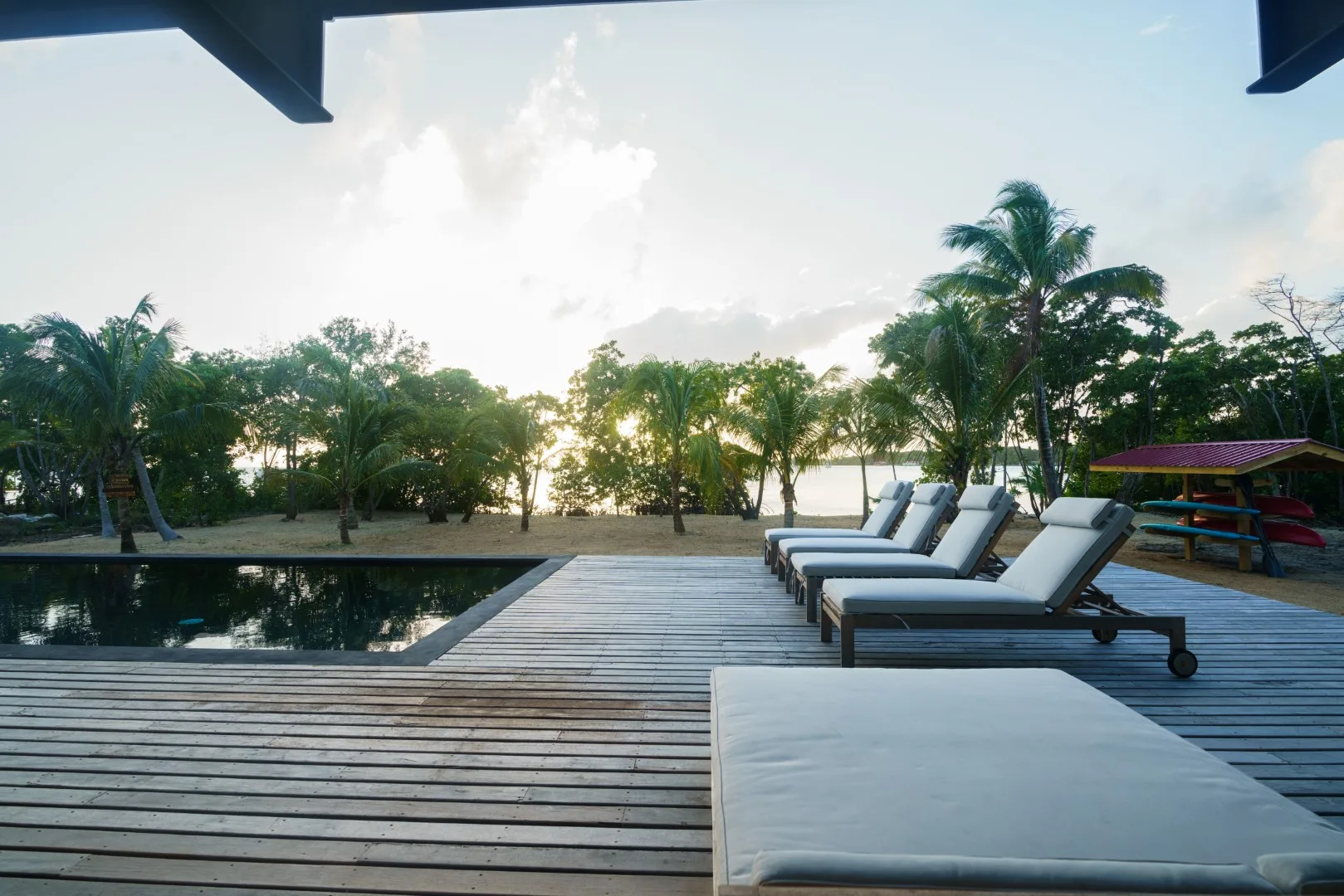 Una terraza de madera junto a una piscina tiene tumbonas acolchadas y vista a los árboles.