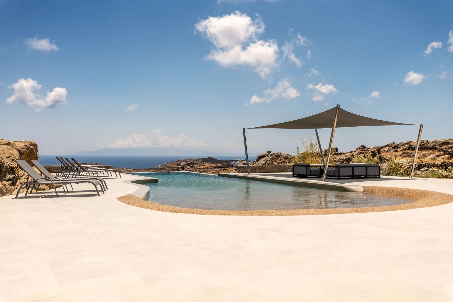 An outdoor swimming pool sits beside lounge seating, with the sea visible beyond.