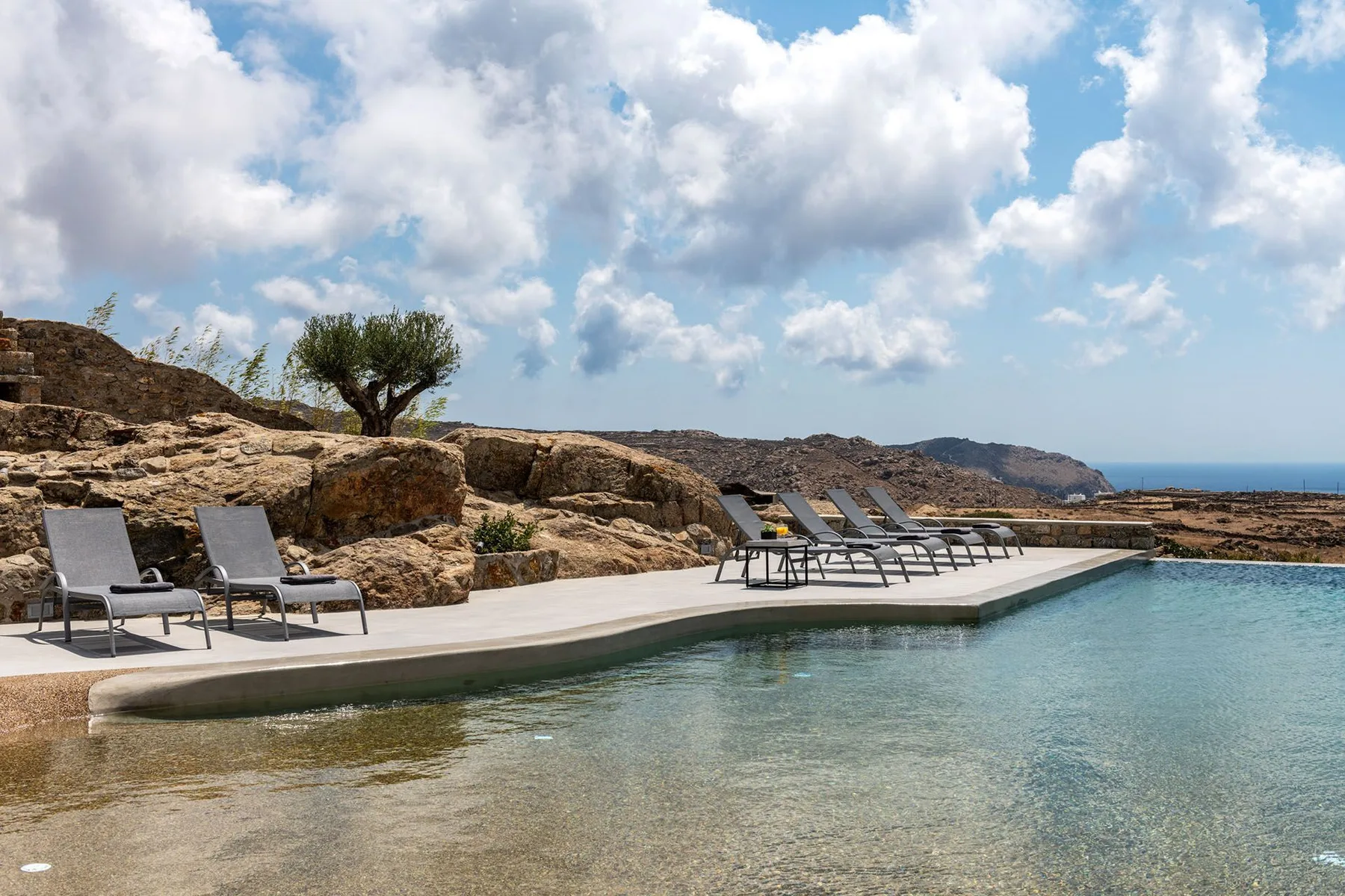 An outdoor swimming pool with lounge seating beside rocky terrain and a distant sea view.
