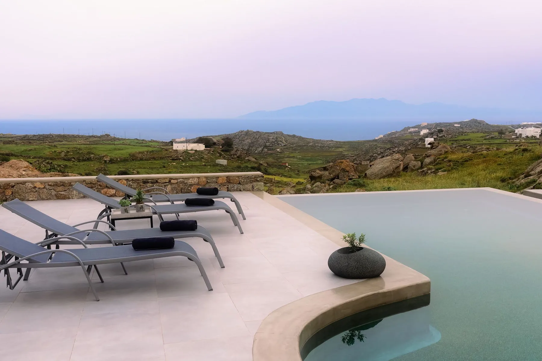 A poolside terrace overlooks green hills and the sea, with loungers beside the water.