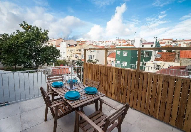 A rooftop terrace has a wooden dining table set with blue dishes overlooking nearby buildings.