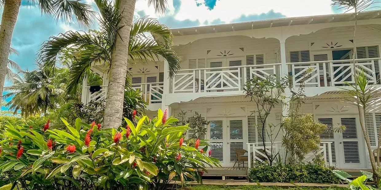 Espacio representativo del hotel: Un edificio blanco de dos pisos con balcones se alza detrás de plantas tropicales.