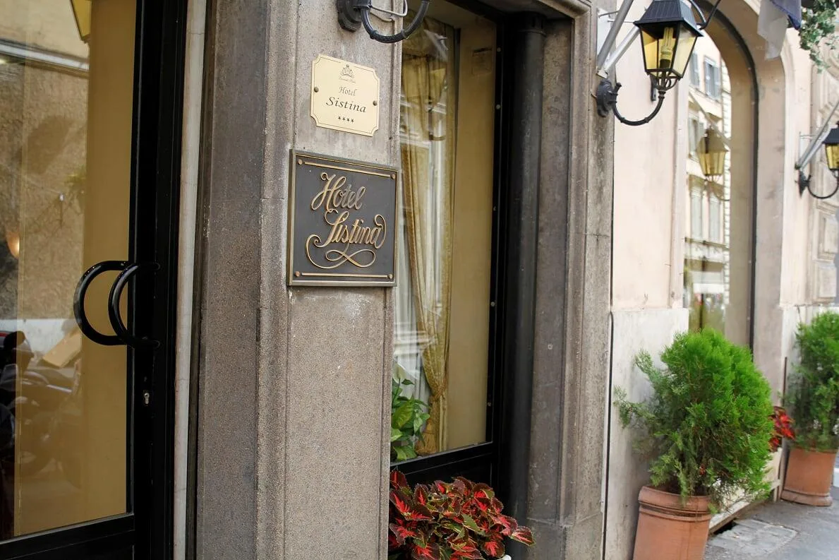 Espacio representativo del hotel: Entrada de hotel con una puerta de vidrio, letreros en la pared y plantas en macetas.