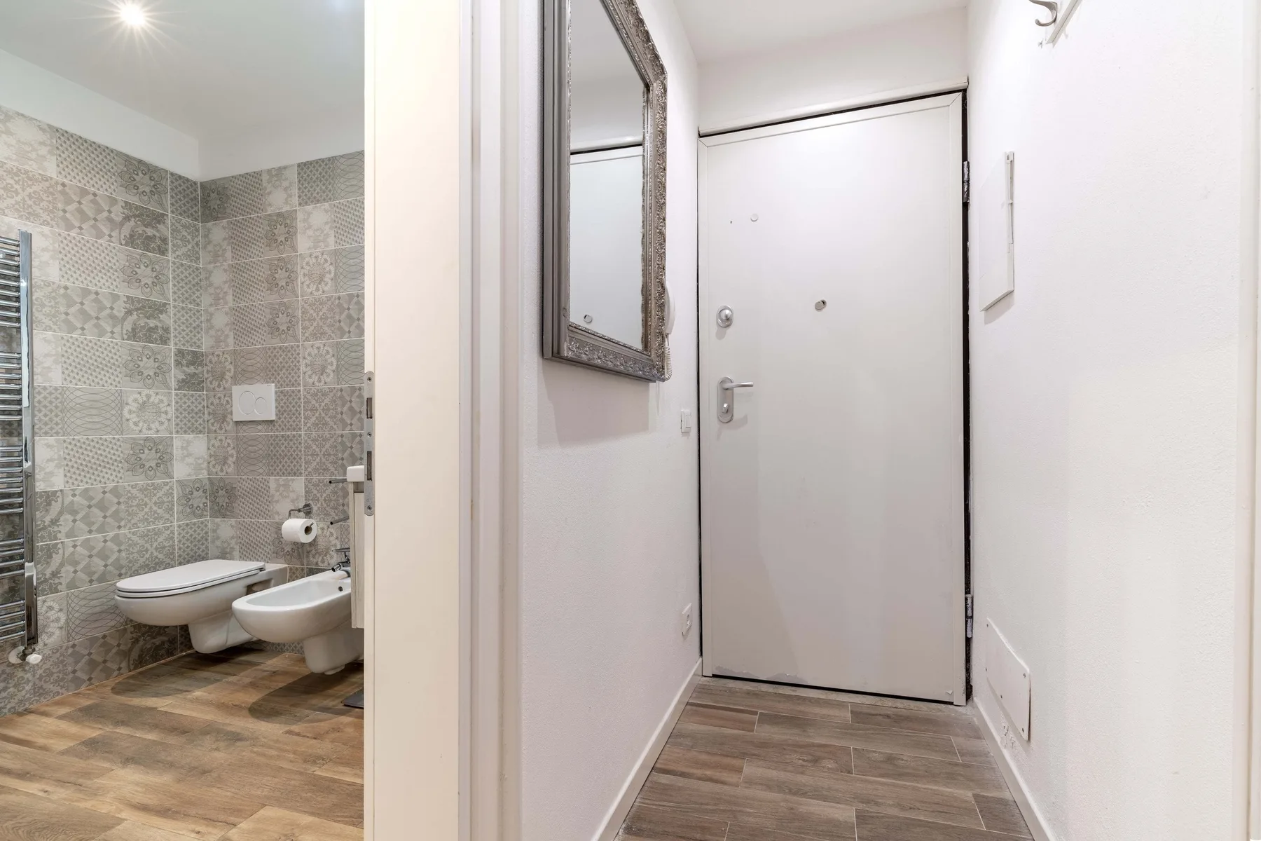 Un pasillo de entrada estrecho con una puerta cerrada, junto a un baño abierto.