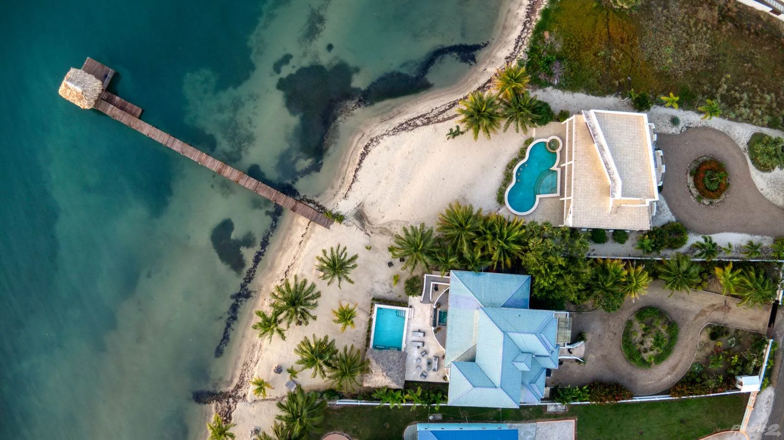 An aerial view shows a beachfront property with a pier, palm trees, and pools.