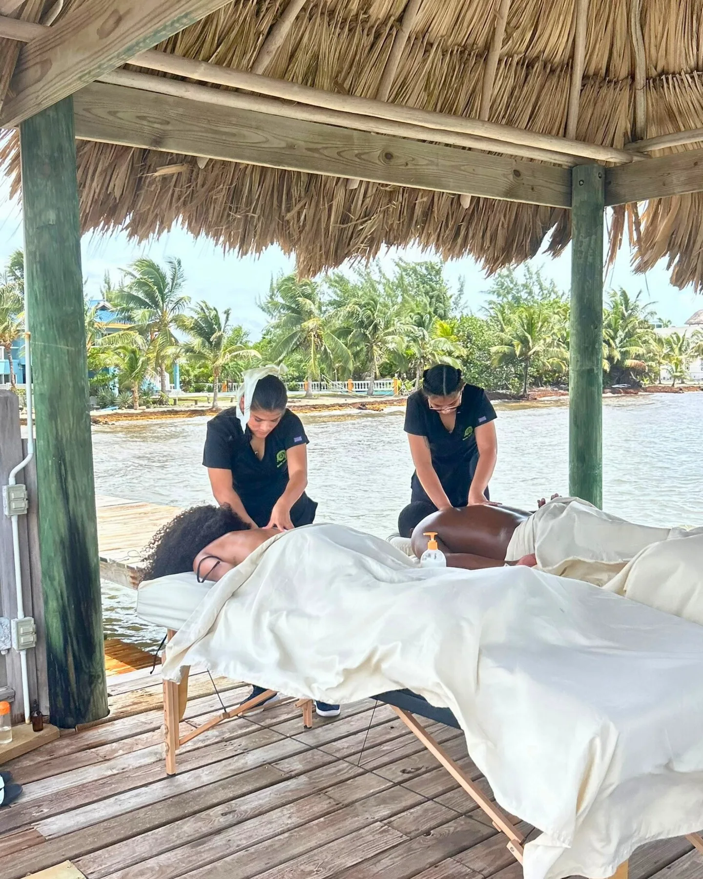 Two people provide a massage beside the water under a thatched shelter.