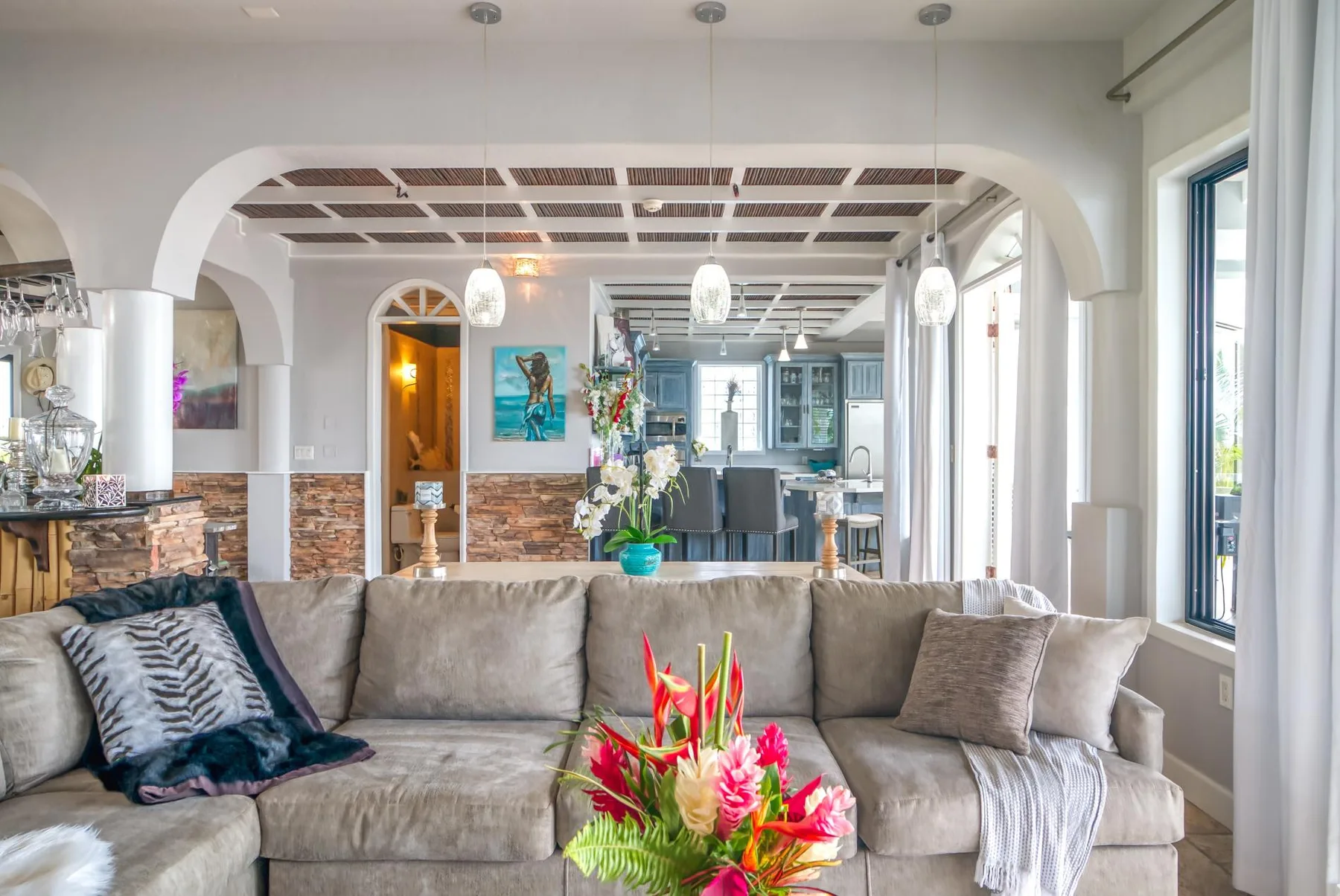 A living room with a sectional seating area, colorful flowers, and an open kitchen beyond.