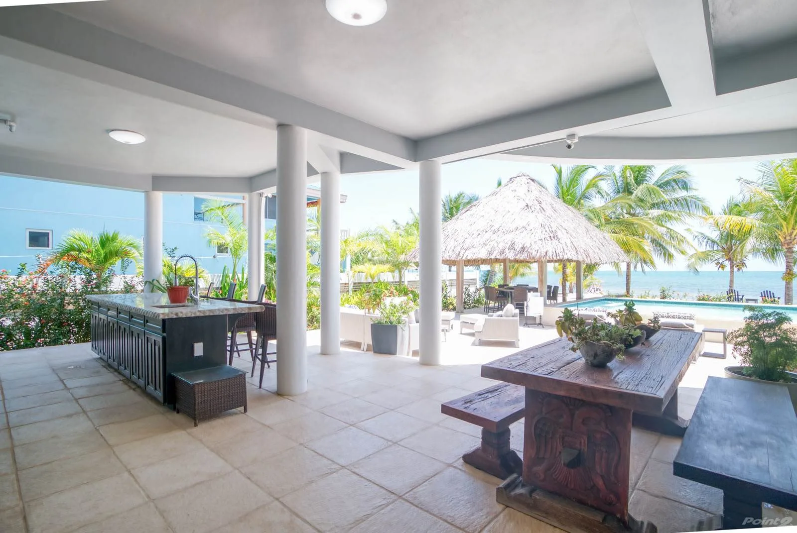 A covered outdoor dining area overlooks a pool, palm trees, and the ocean.