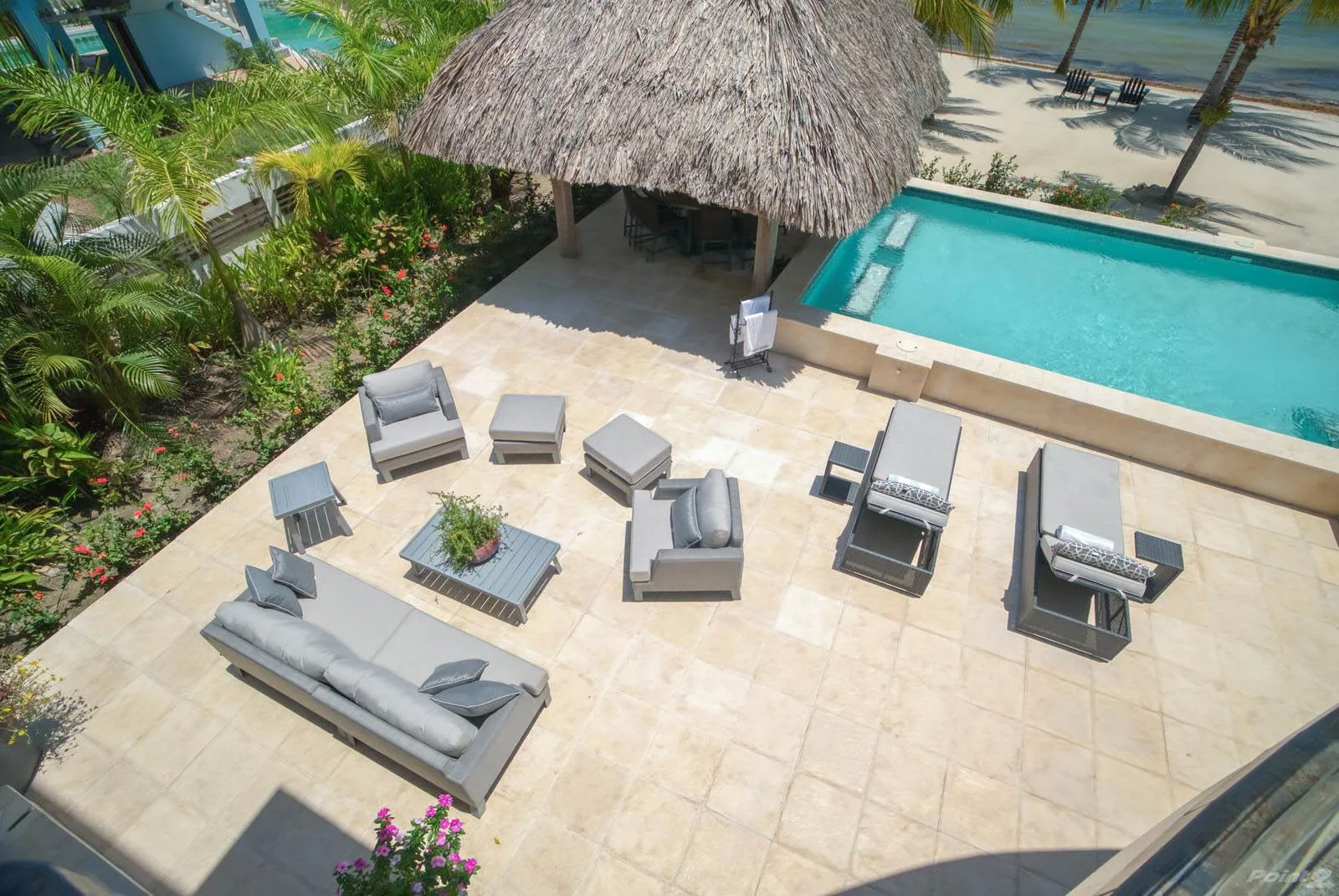 A tiled poolside patio has lounge seating beside a thatched-roof pavilion.