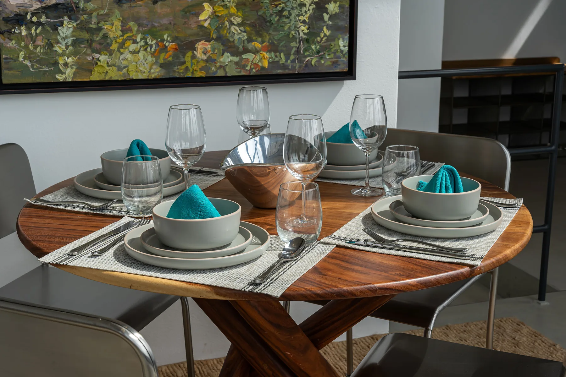 A dining table is set with plates, glassware, and teal napkins beneath a framed painting.