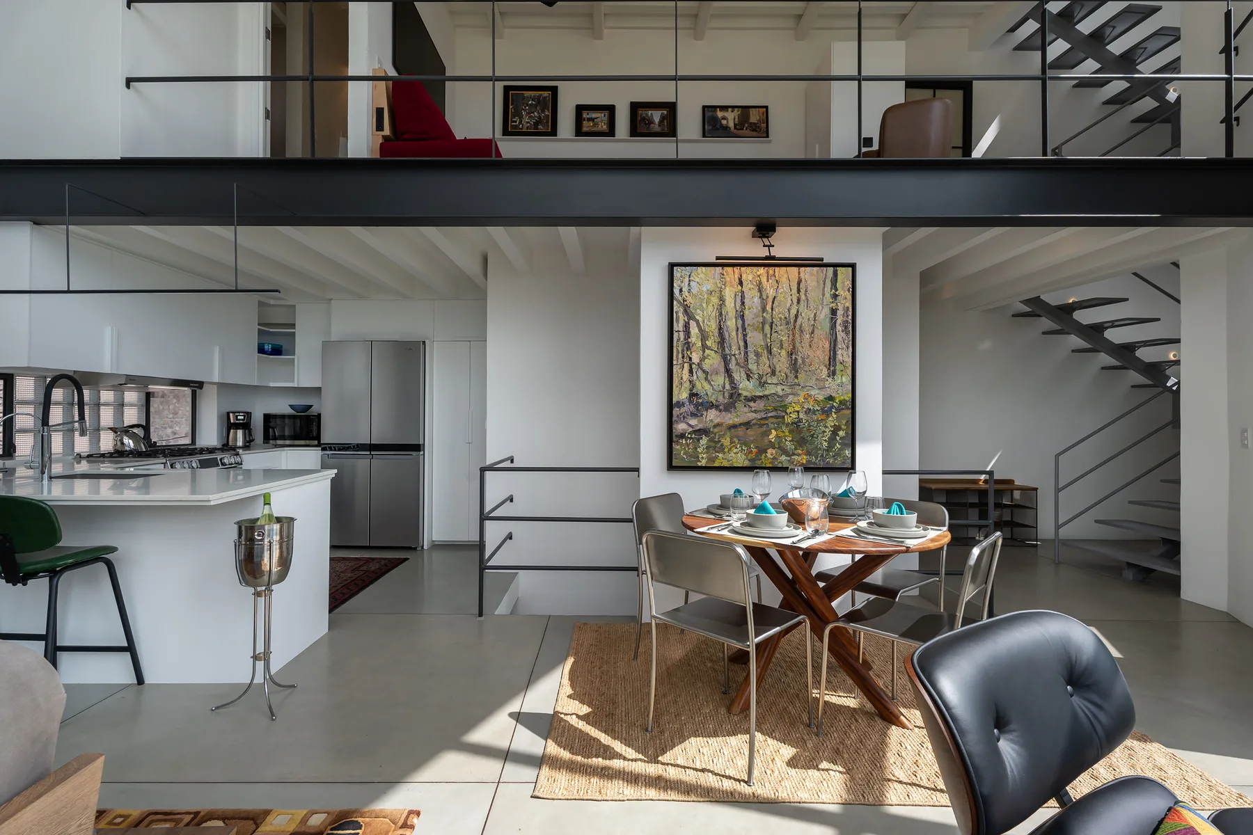 An open-plan dining and kitchen area with a table beneath a large forest painting.