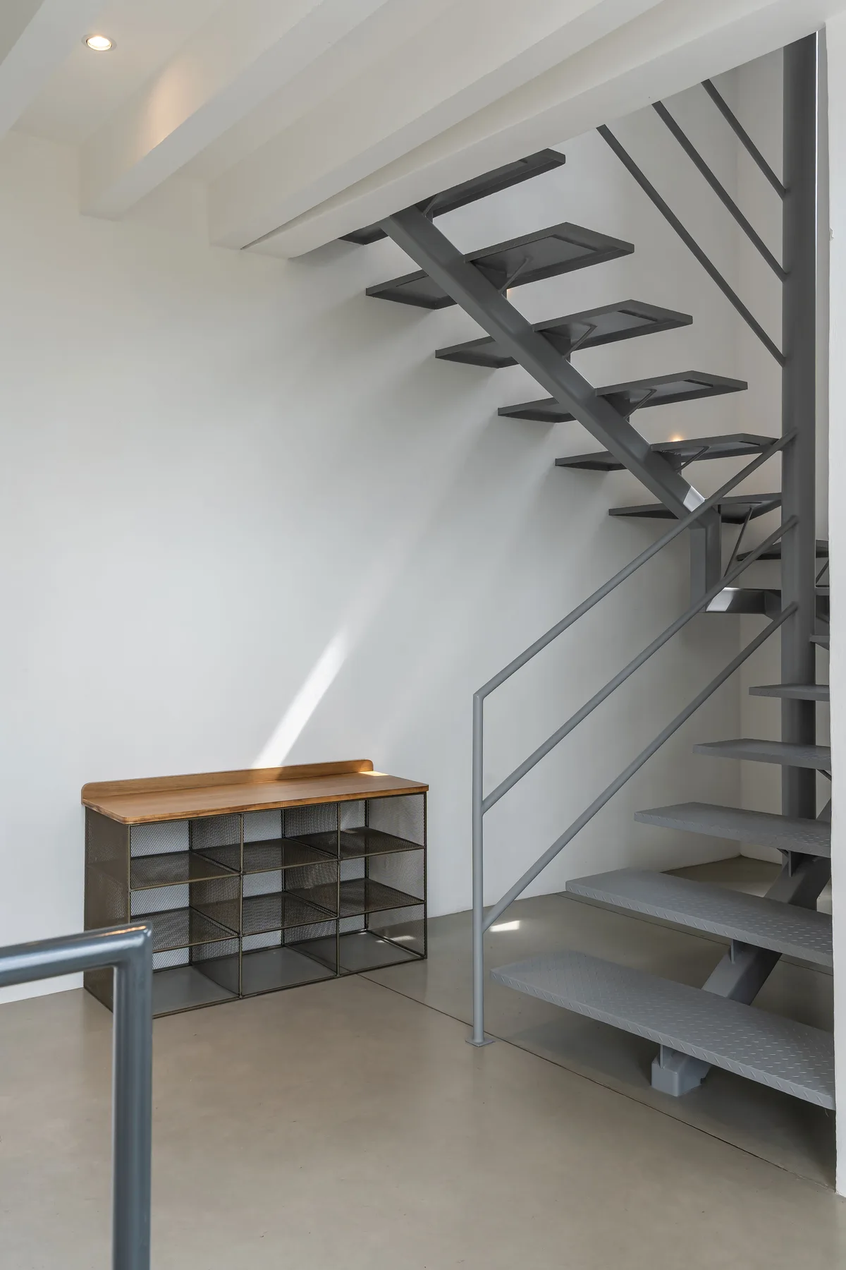 A modern staircase rises beside a low shelving unit in a bright interior.