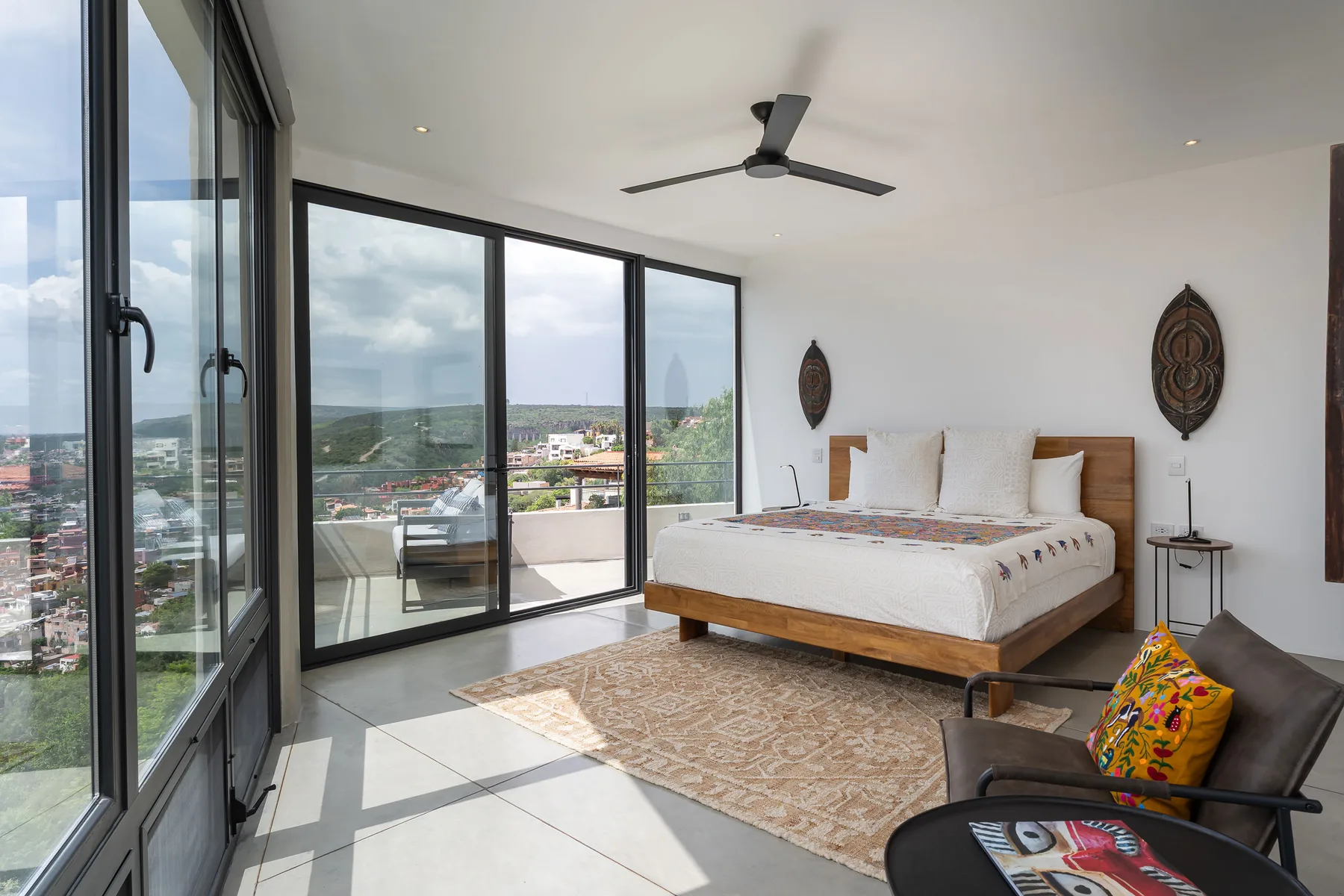 A bright bedroom with a bed beside floor-to-ceiling windows overlooking the city.