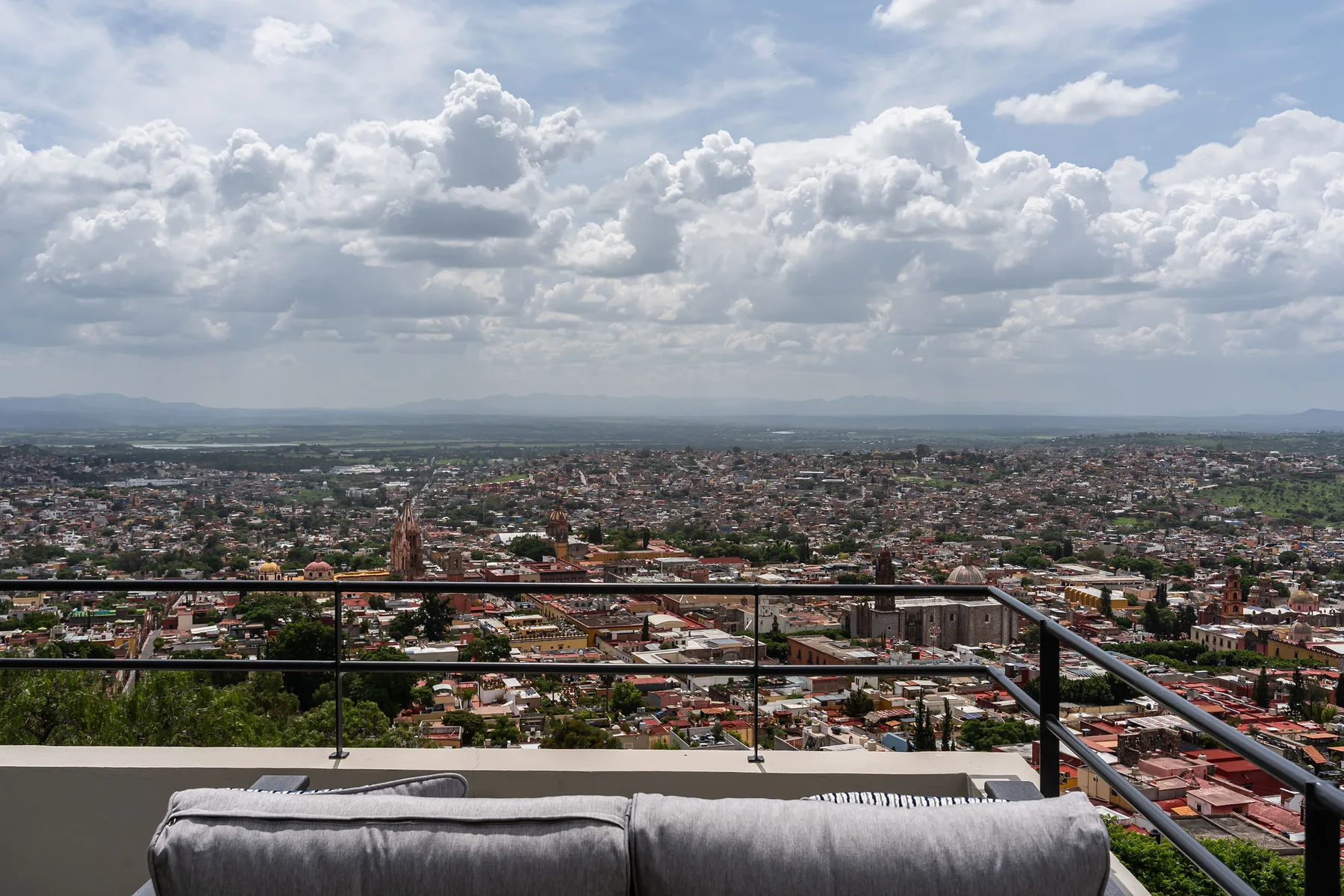 A cityscape stretches toward distant hills beneath a cloudy sky, viewed from a terrace.