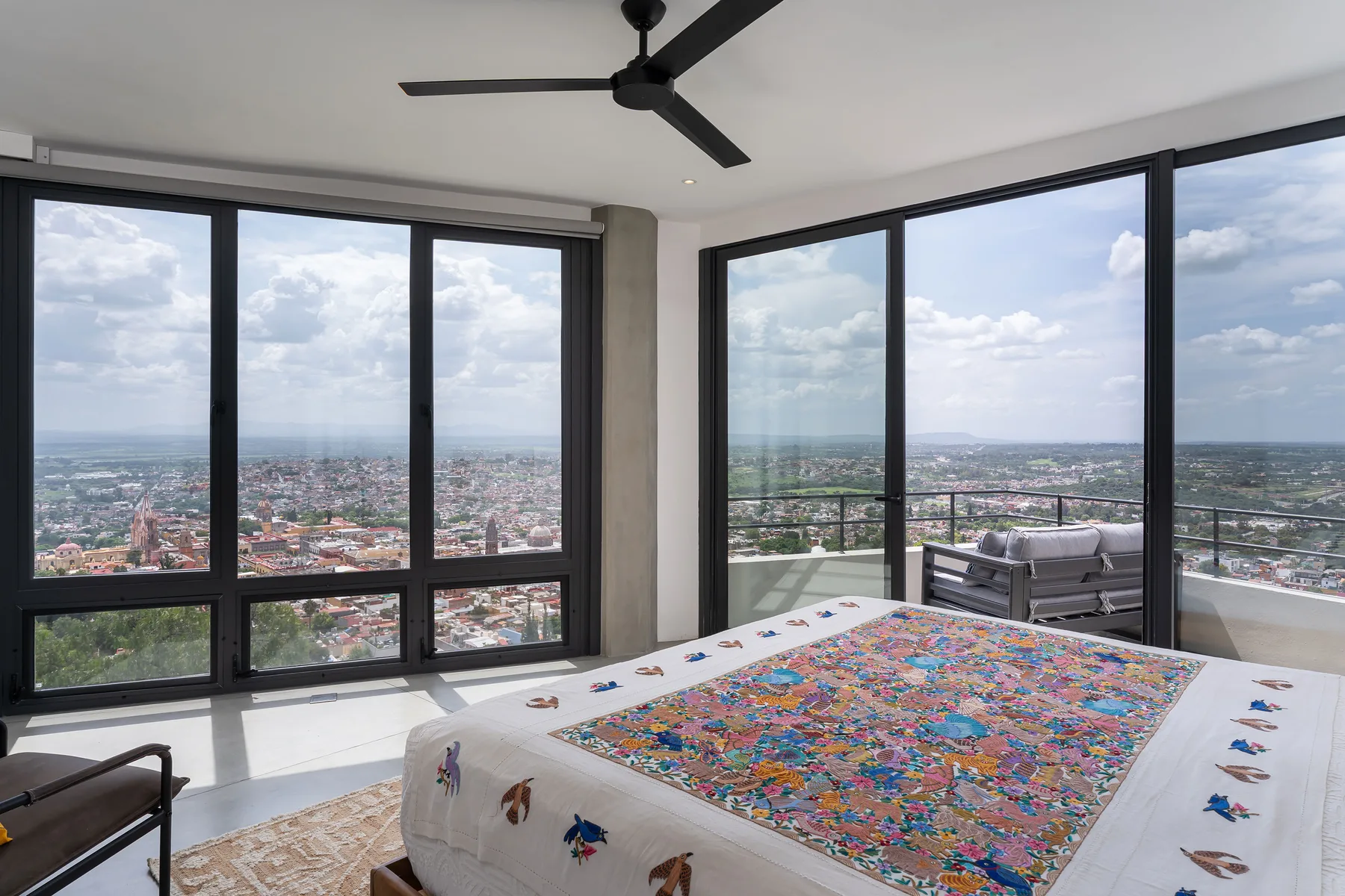 A bedroom with a large bed beside floor-to-ceiling windows overlooking the city.