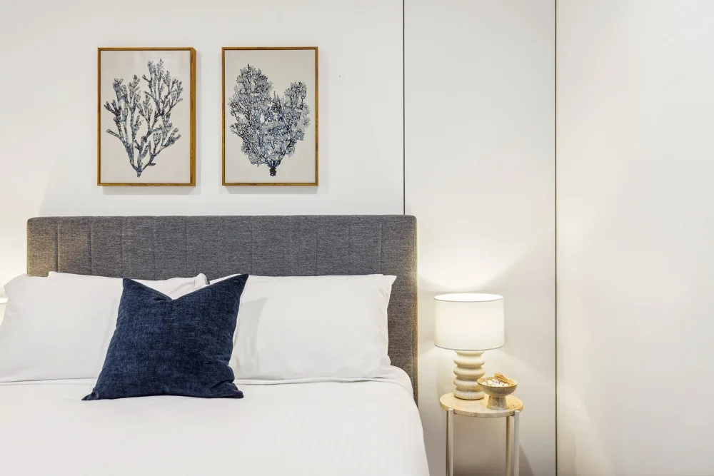 A bedroom with a neatly made bed, dark accent pillow, and framed botanical prints.