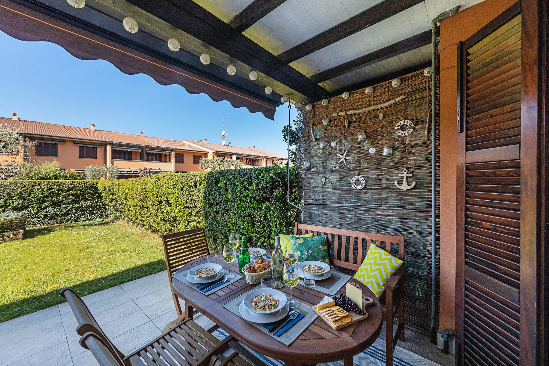 A covered patio has a dining table beside a garden hedge.
