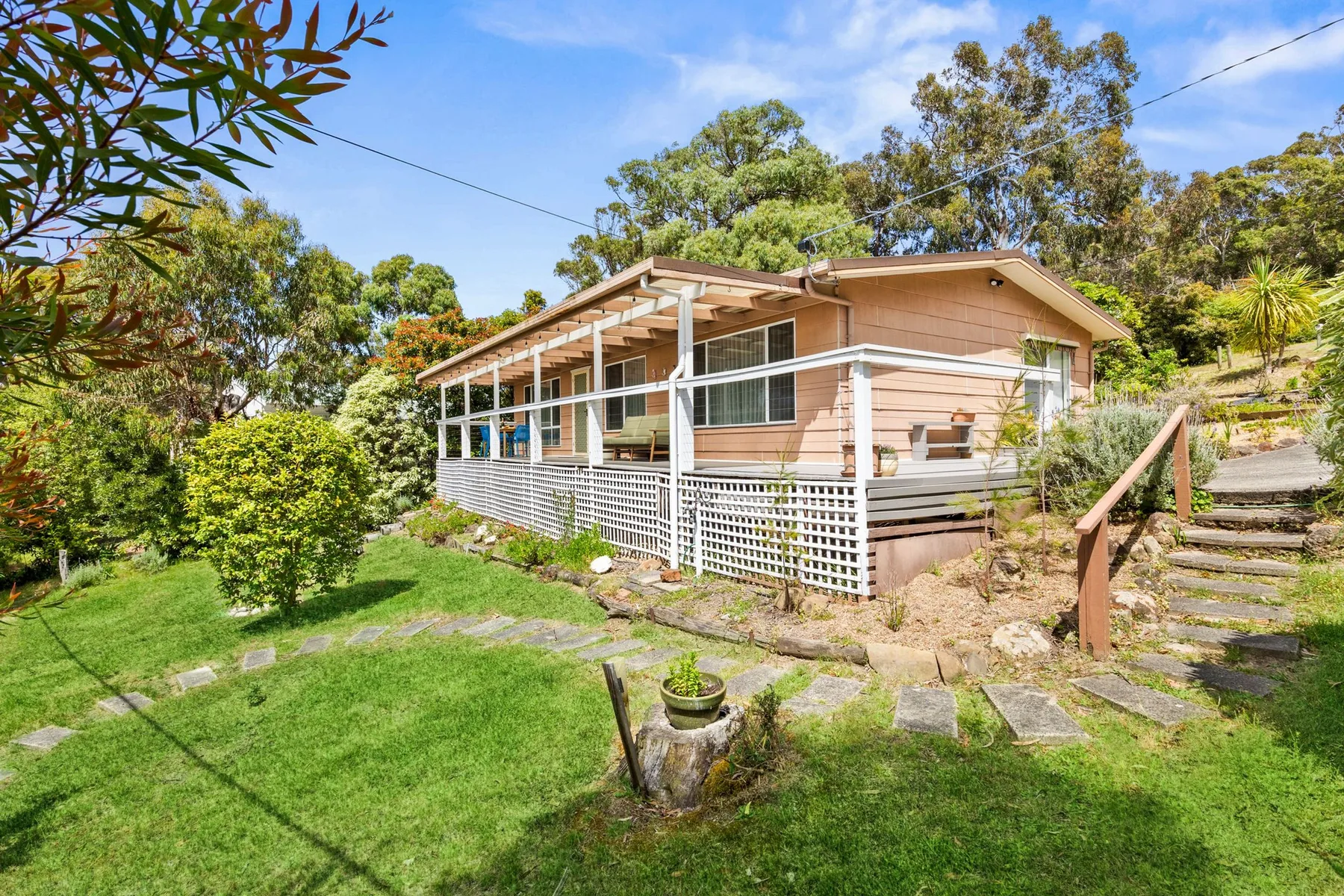 A raised house with a covered porch sits beside a grassy garden and stone steps.