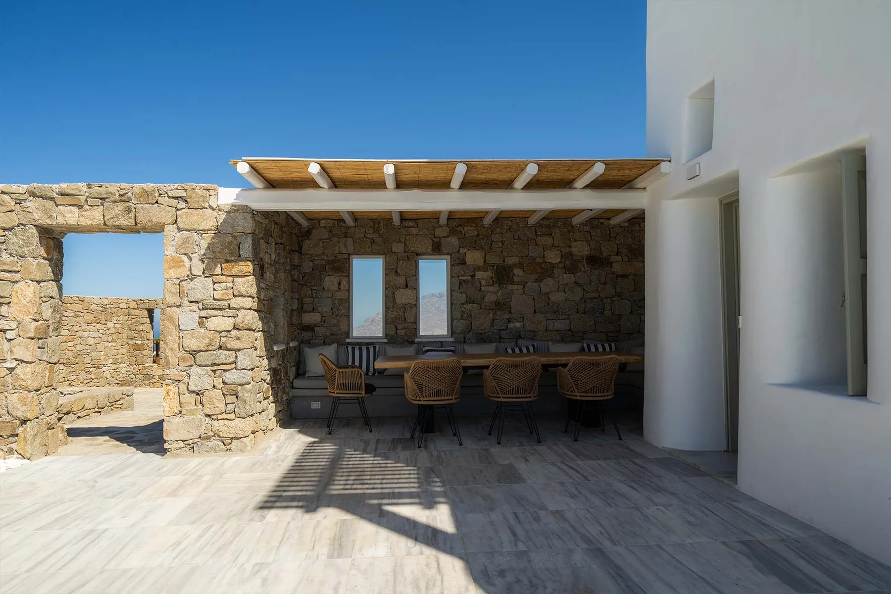 A shaded outdoor dining area with a long table and woven seating beside a stone wall.