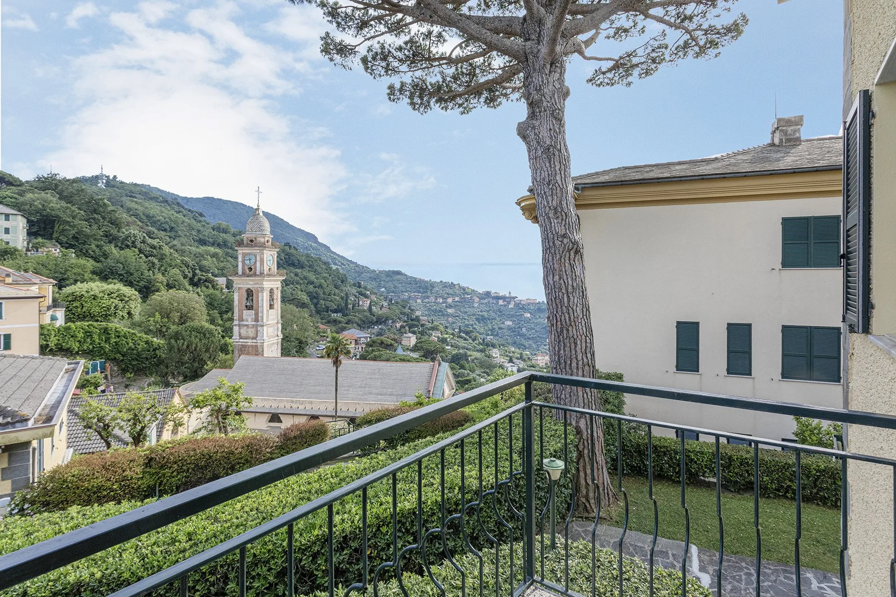 A balcony overlooks a hillside town, with a bell tower and a large tree in view.