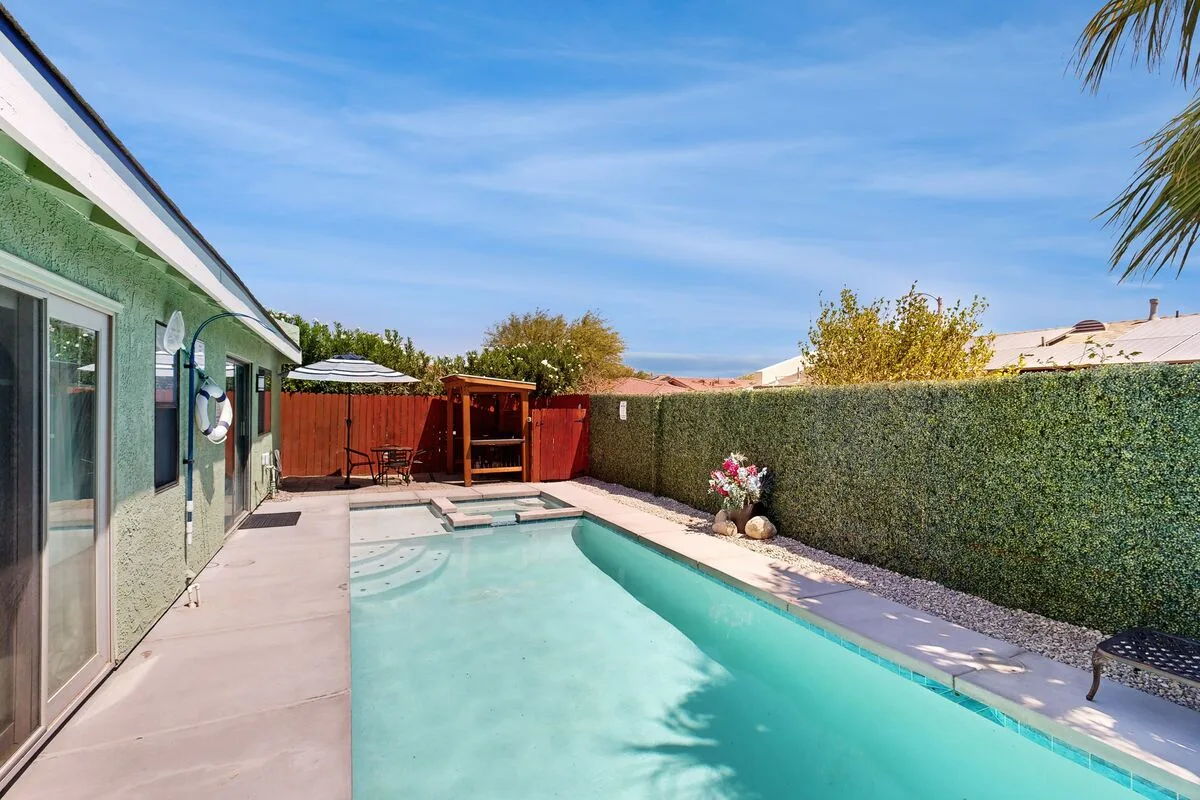 Una piscina al aire libre junto a una casa, con una terraza pavimentada y un seto.