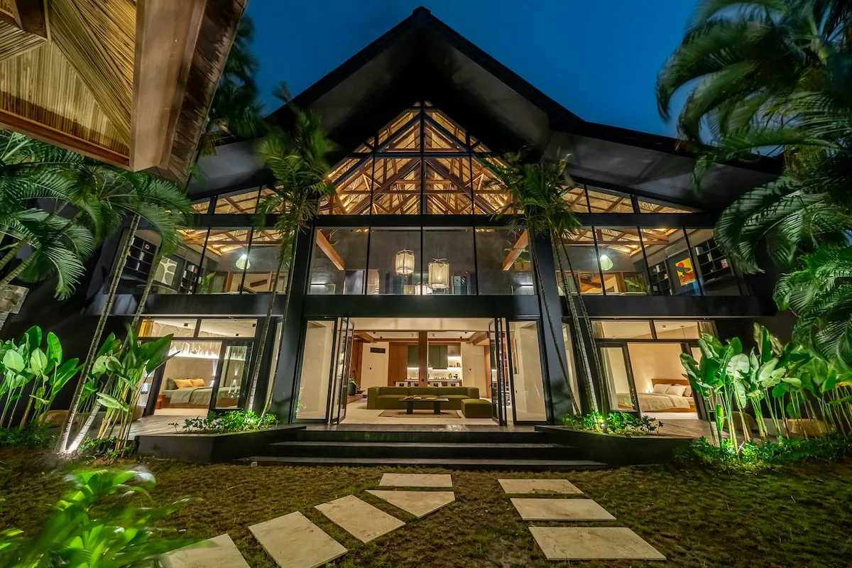 A large glass-fronted building is illuminated among tropical plants at night.