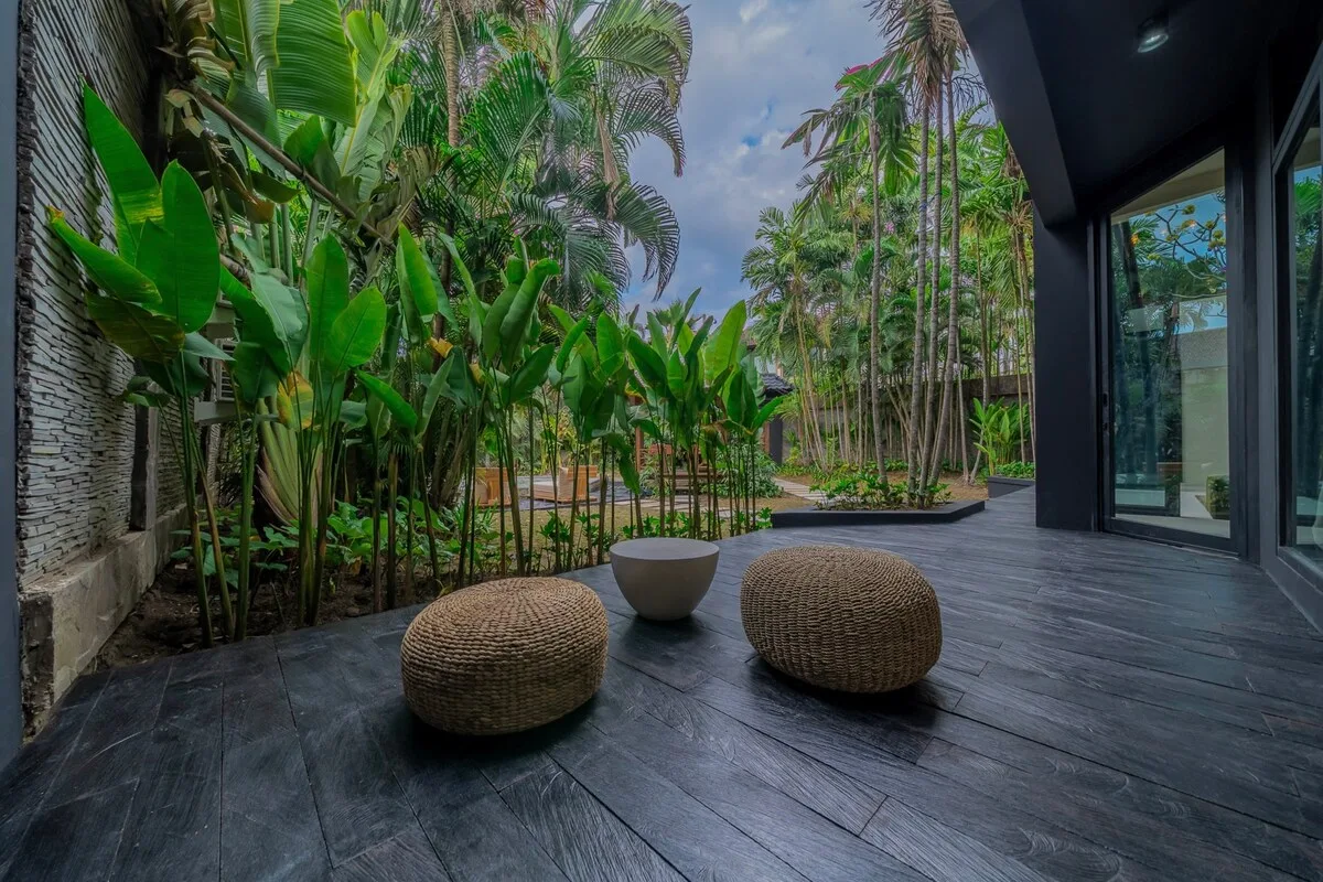 A tropical garden patio with leafy plants, woven seating, and a dark tiled floor.
