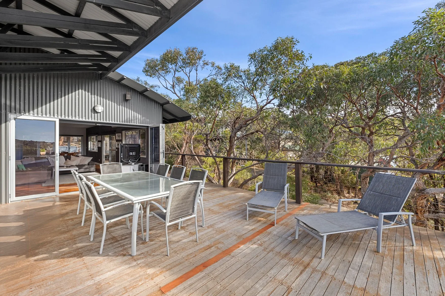 A covered wooden deck with a glass dining table and outdoor seating overlooks leafy trees.