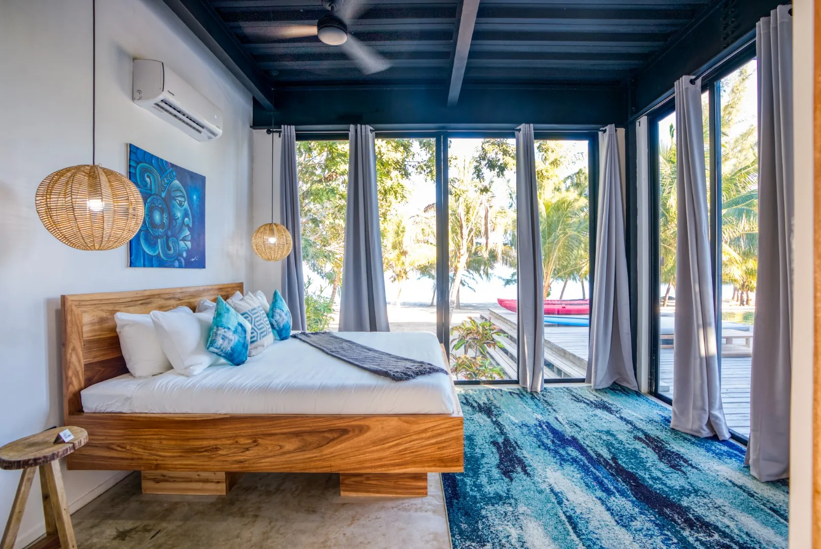A bedroom with a wooden bed, blue rug, and large windows overlooking palm trees.