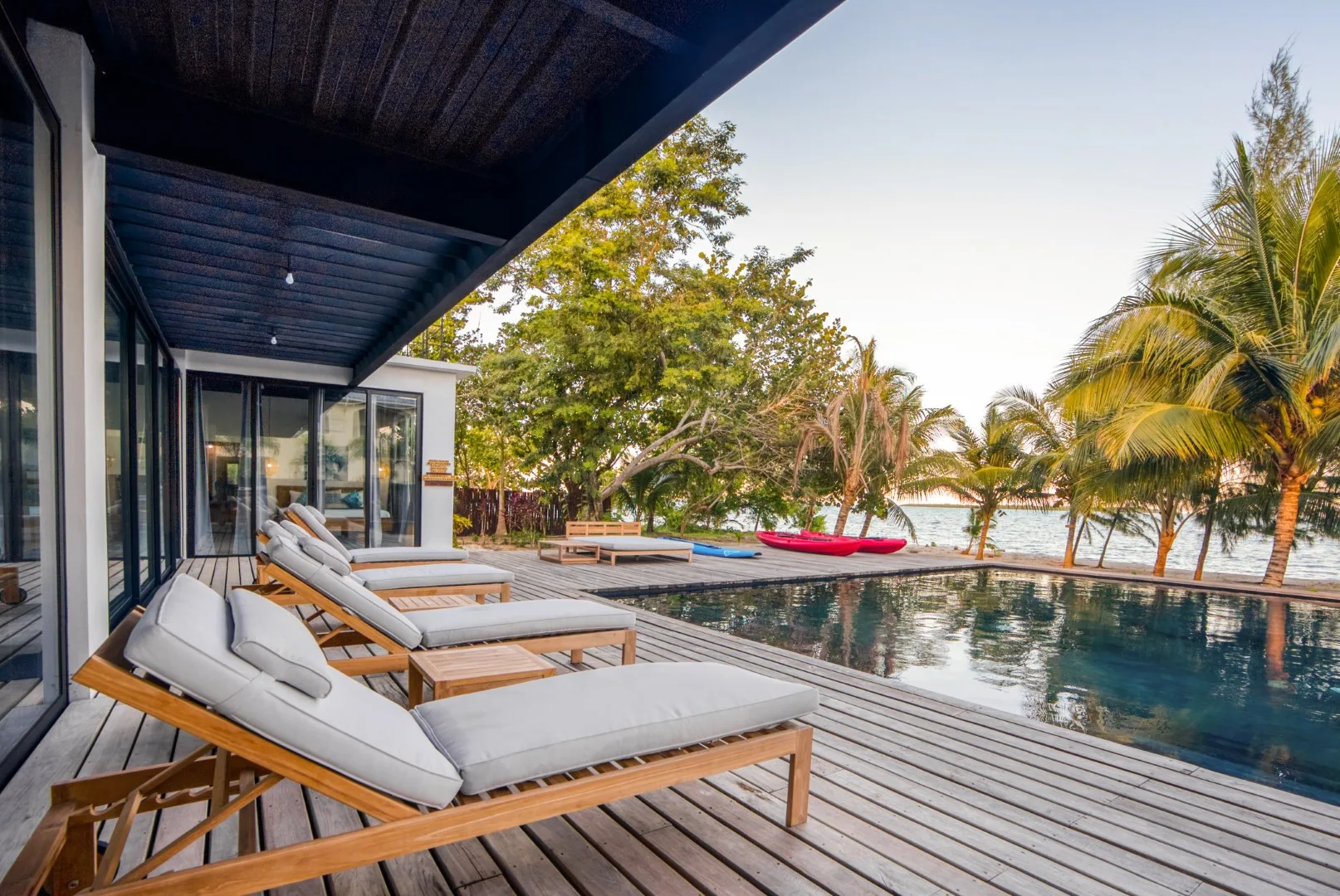 Poolside loungers sit on a wooden deck beside a modern house, with palm trees beyond.