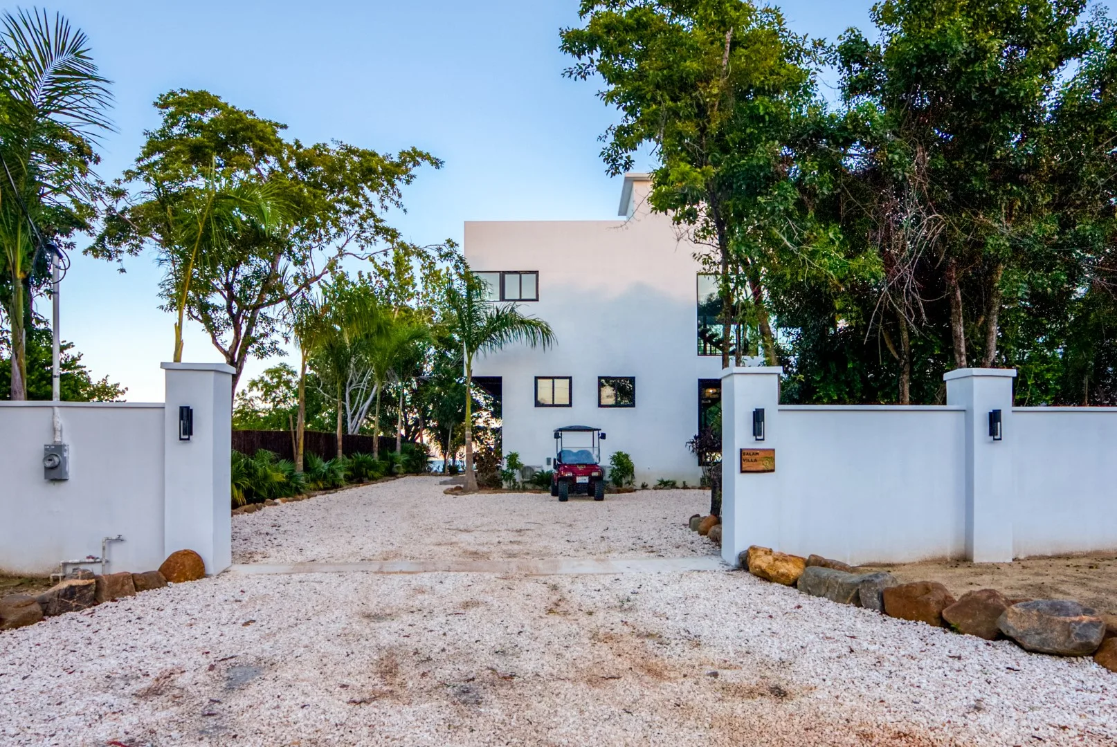 A white building stands beyond a gravel driveway, framed by trees and gate walls.
