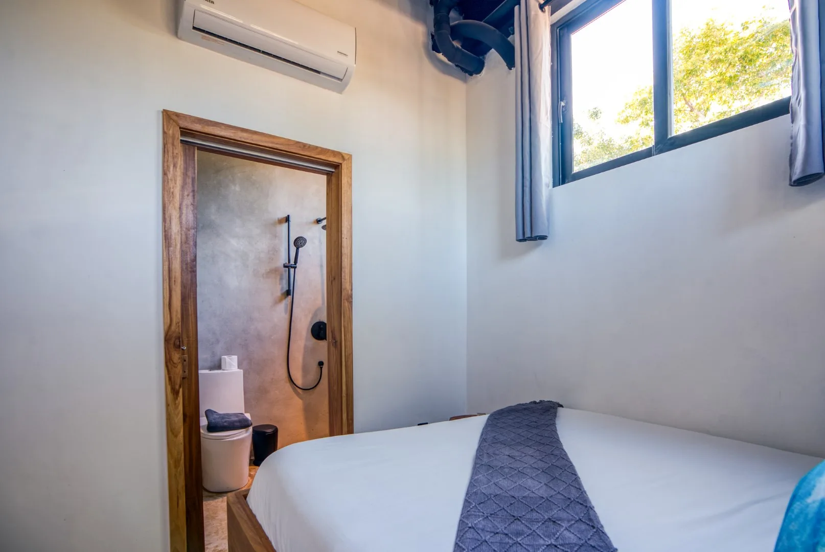 A bedroom with a neatly made bed beside a window and an adjoining bathroom.