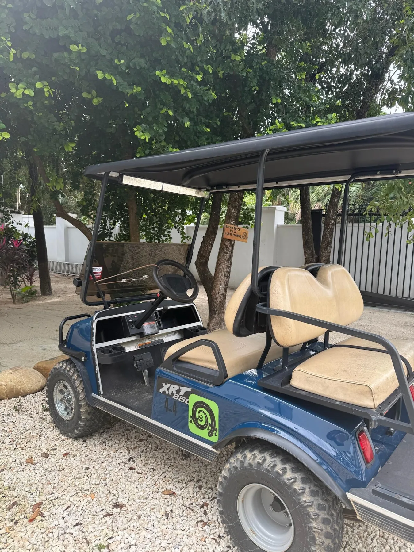 A blue golf cart with a roof is parked beneath leafy trees on a gravel path.