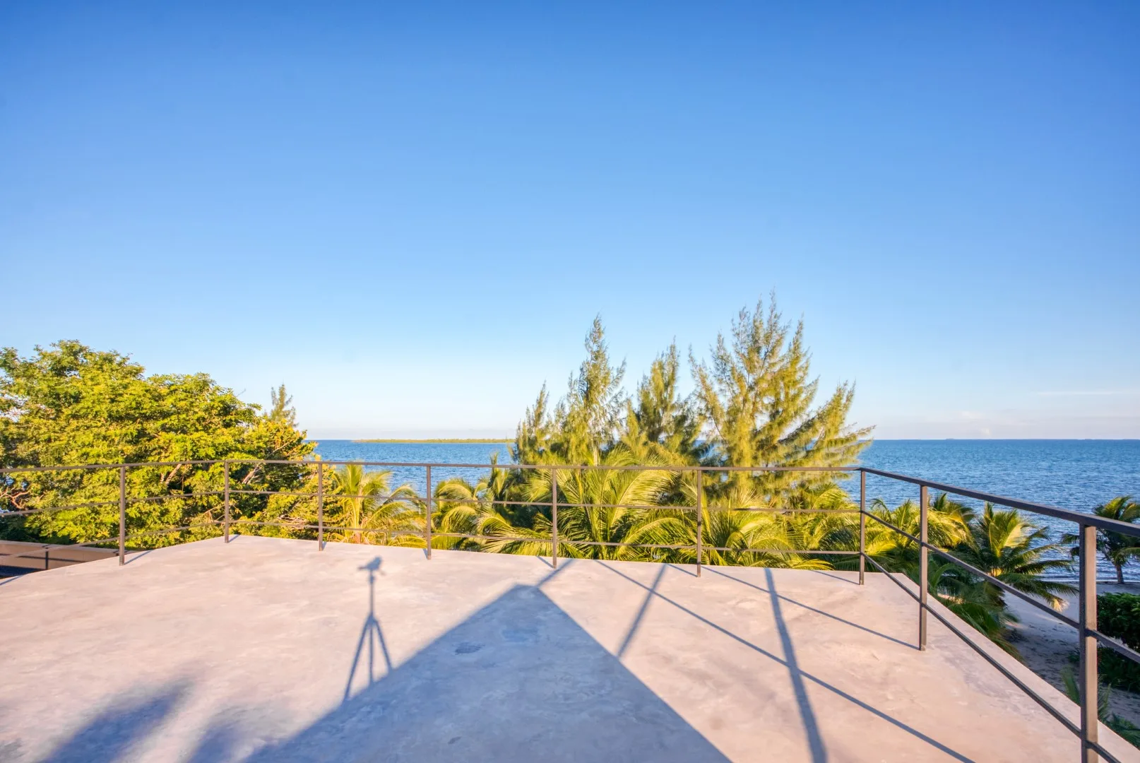 A sunny oceanfront terrace overlooks the water, with trees along the railing.