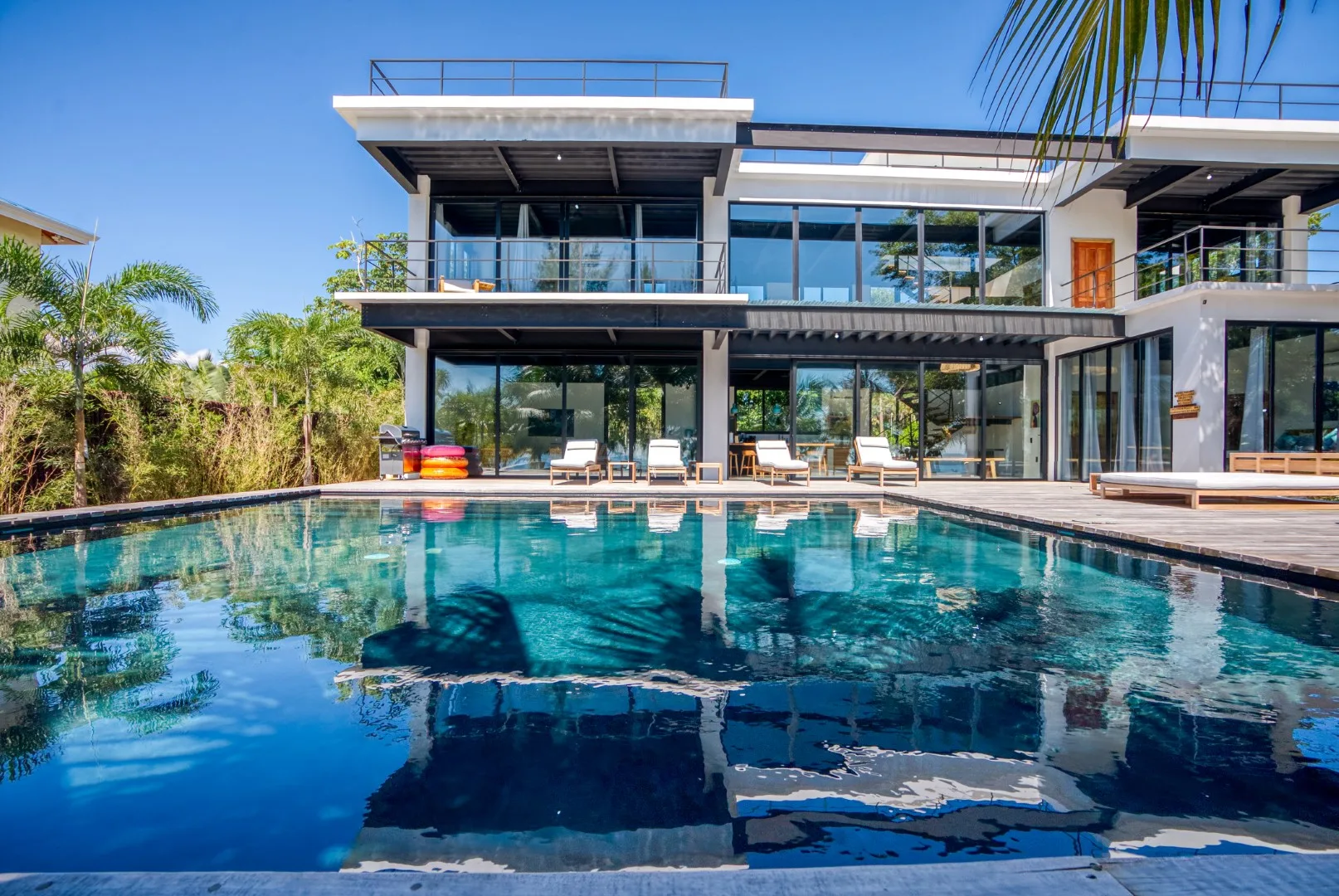 A modern glass-fronted house overlooks a swimming pool with lounge seating.