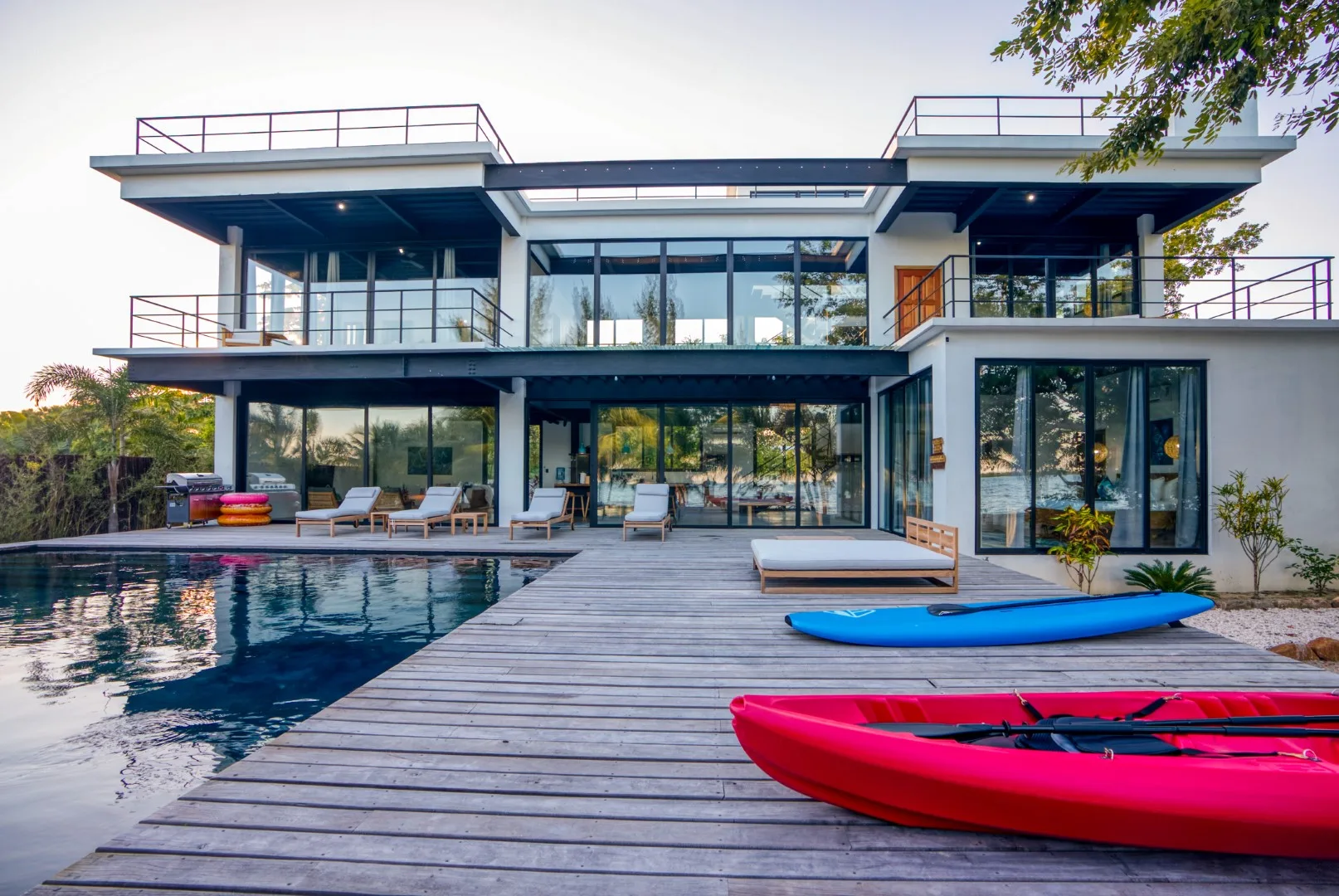 A modern glass-fronted house stands beside a pool and wooden deck.