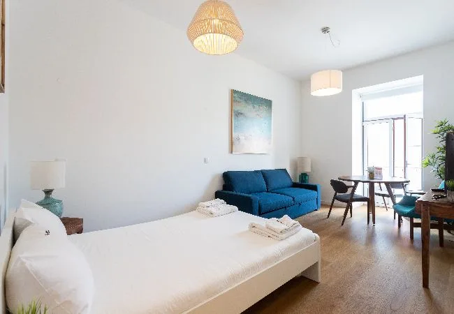 Una habitación luminosa combina una cama, asientos azules y una mesa de comedor junto a la ventana.